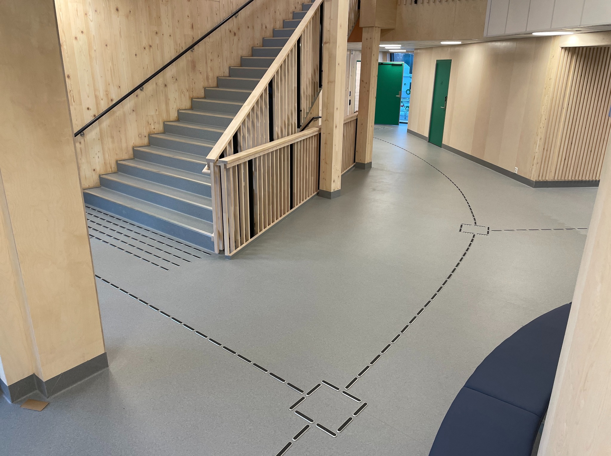 Szara posadzka żywiczna z dekoracyjnymi elementami w kształcie kwadratów i linii, drewniane schody i ściany, nowoczesny design wnętrza budynku użyteczności publicznej.