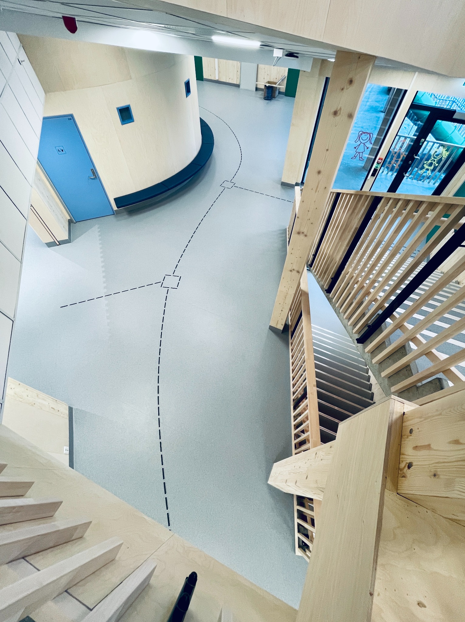 Widok z góry na nowoczesne wnętrze z drewnianymi schodami, szarą posadzką z czarnymi liniami i niebieskimi drzwiami. Jasne oświetlenie, minimalistyczny design.