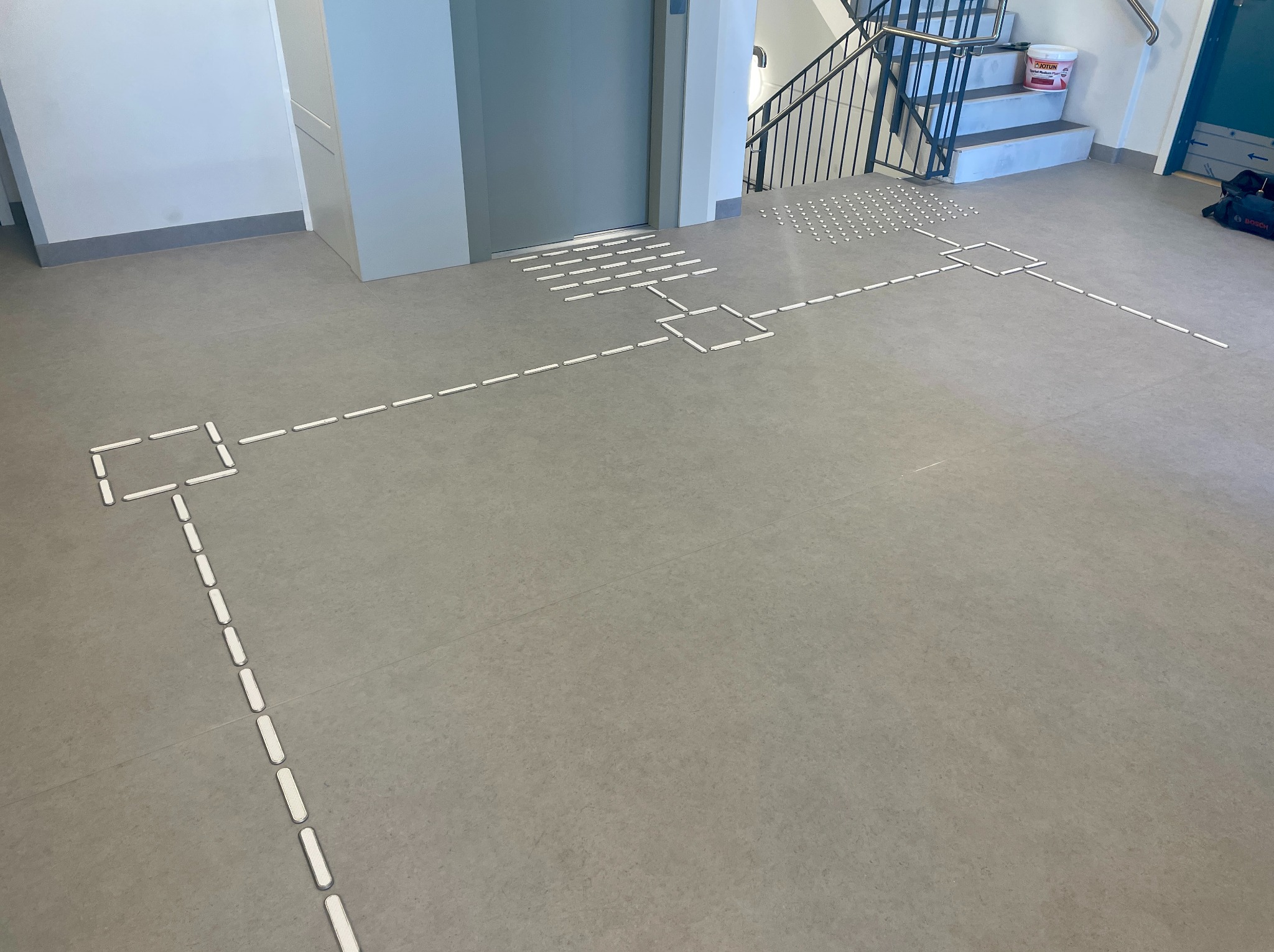 Szara posadzka z białymi, wypukłymi oznaczeniami dla osób niewidomych, prowadzącymi od windy do schodów. Widoczny fragment narzędzi i farby.
