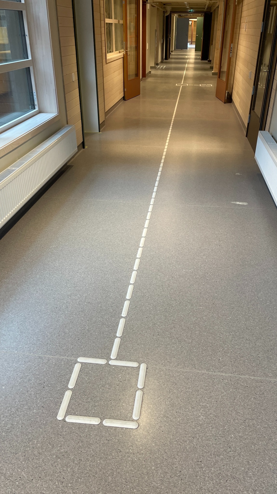 Szara posadzka w korytarzu z białymi, prostokątnymi elementami tworzącymi linię i kwadrat, prawdopodobnie oznaczenia lub dekoracja.