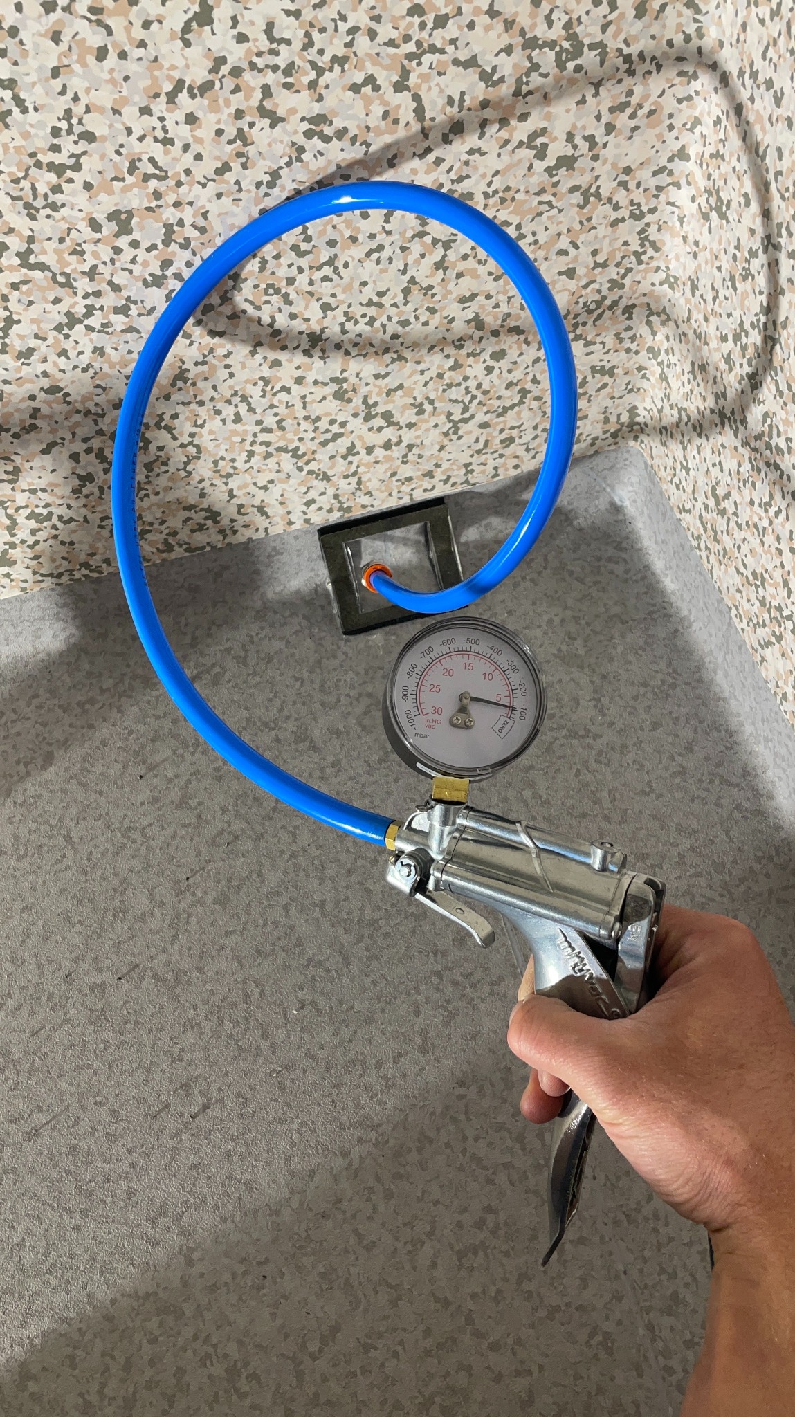 Pistolet do pomiaru ciśnienia w dłoni na tle posadzki w Koronowie. Narzędzie z manometrem i niebieskim przewodem. Kontrola jakości wylewki.