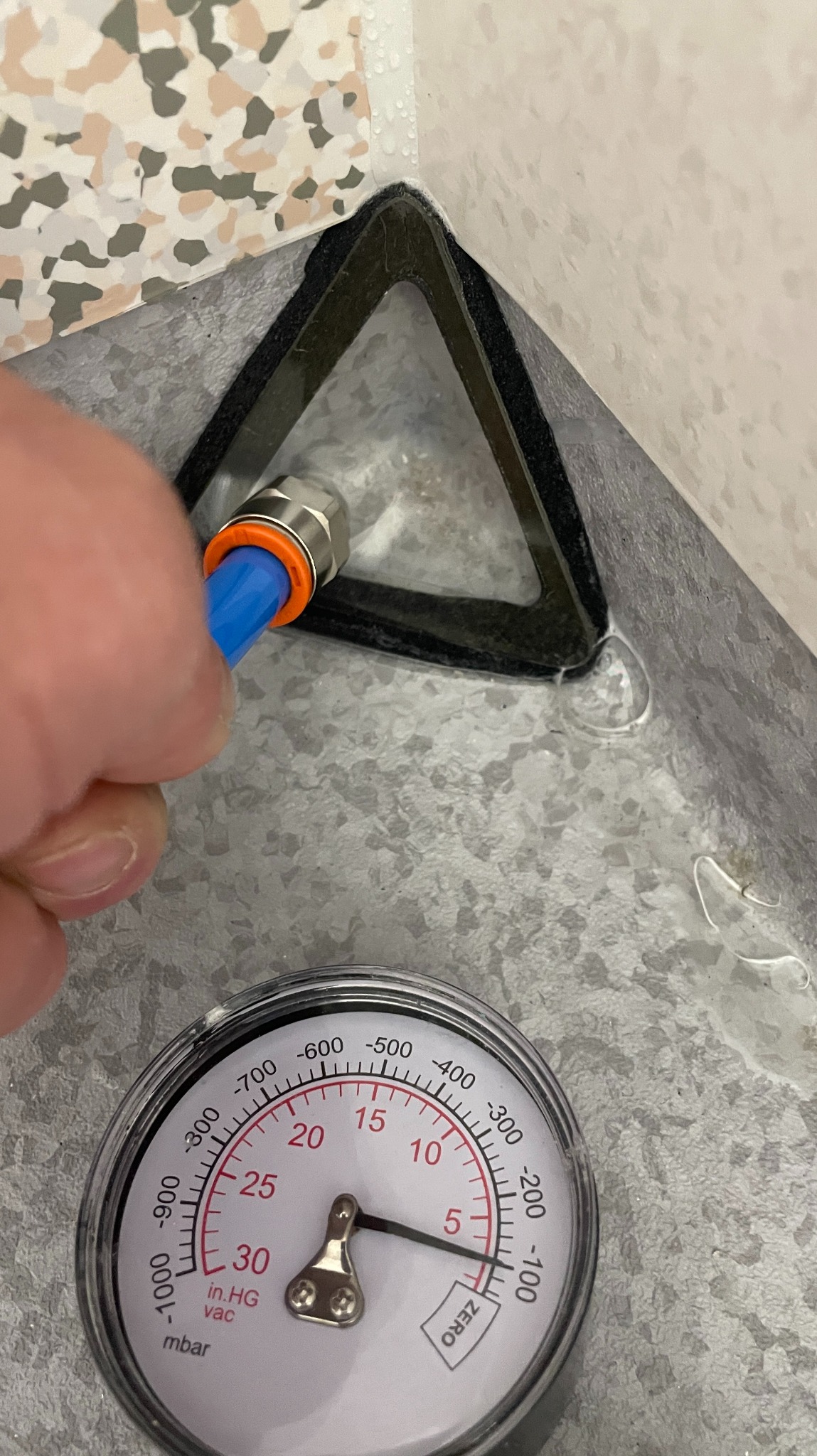 Sprawdzanie szczelności posadzki żywicznej w rogu pomieszczenia za pomocą węża i manometru. Widoczne złącze i czarny uszczelniacz narożny.