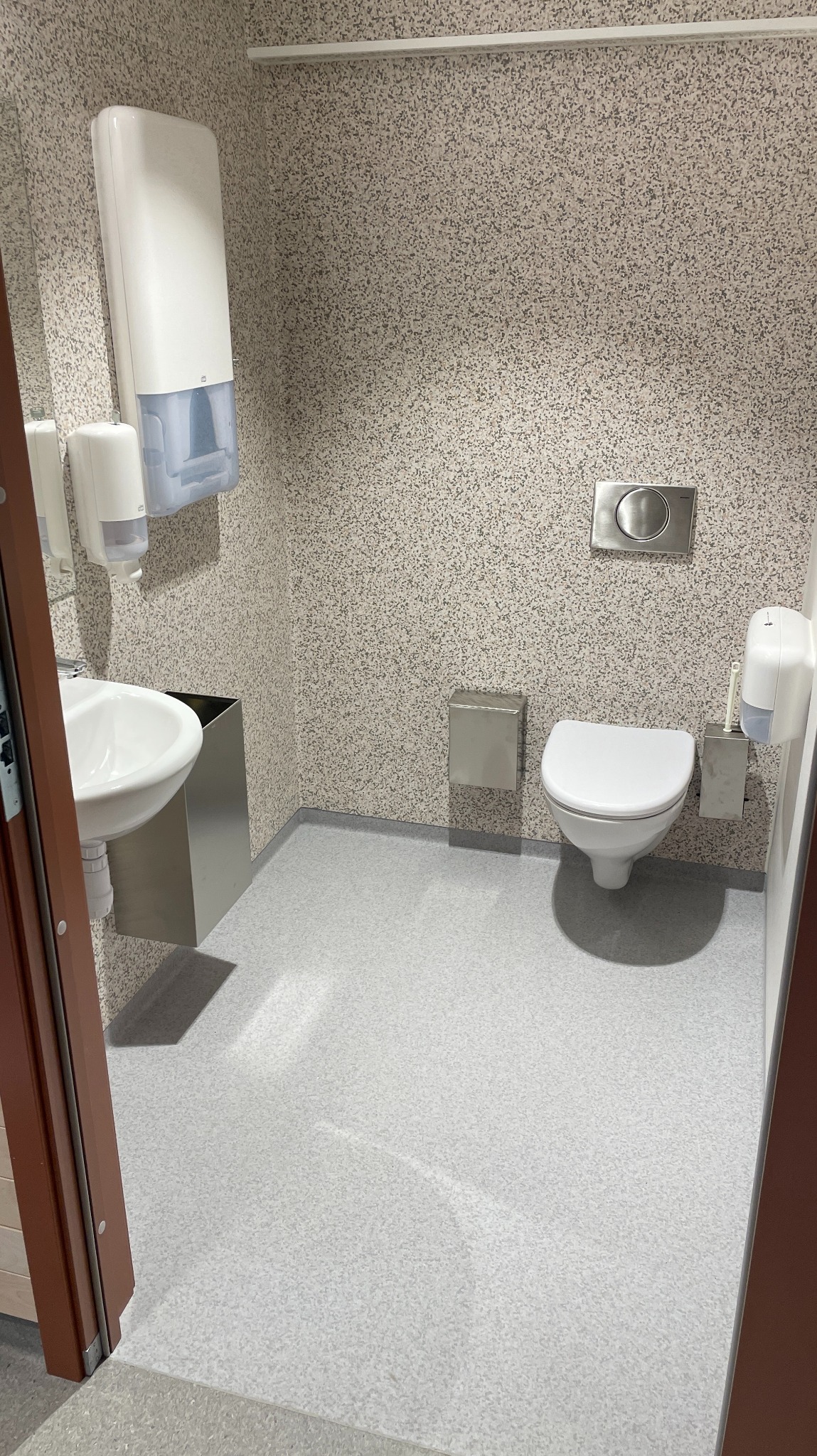 Wnętrze toalety publicznej z szarą posadzką i ścianami w jasny wzór. Widoczna umywalka, dozowniki mydła i papieru, toaleta oraz stalowe akcesoria.