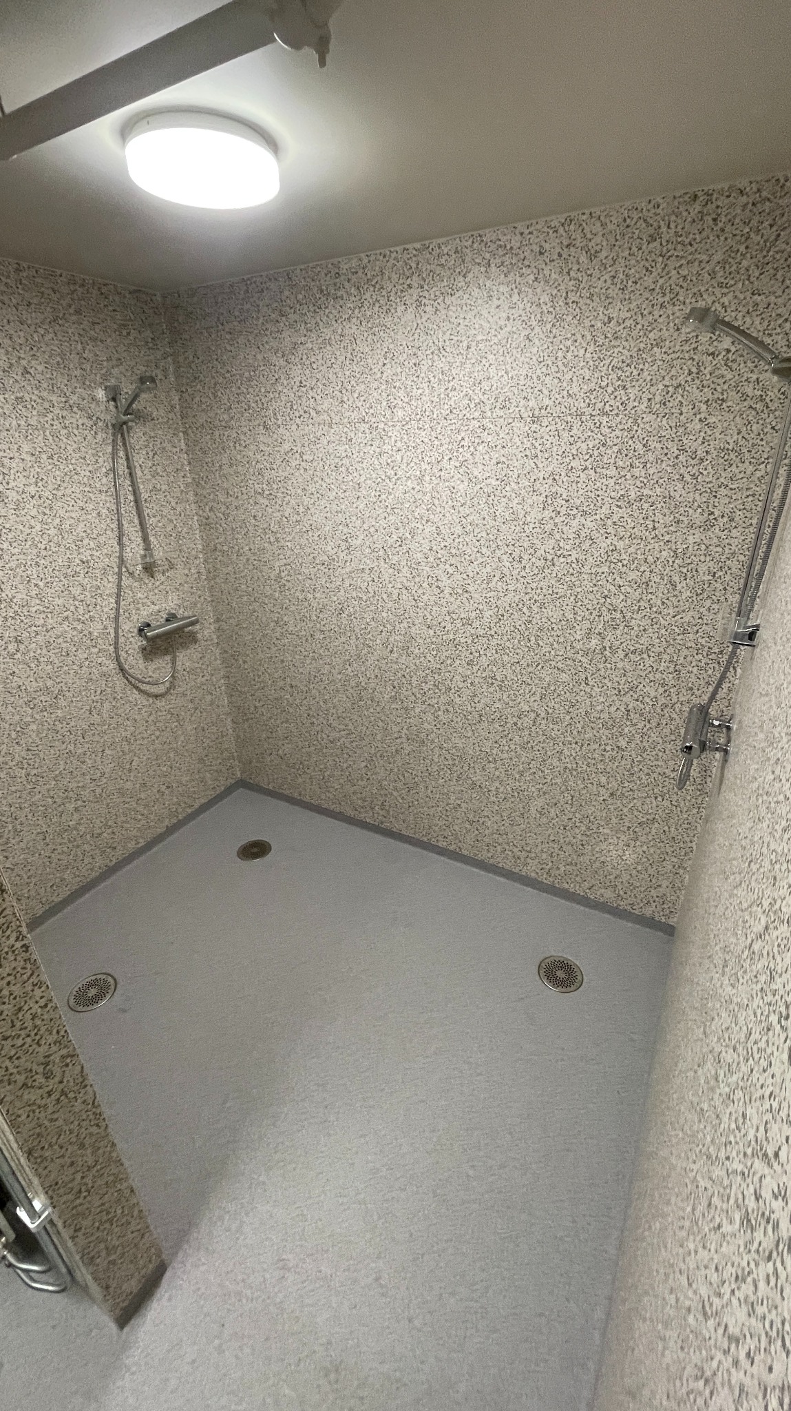 Szara posadzka w kabinie prysznicowej z odpływami i teksturowanymi ścianami. Widoczne baterie prysznicowe.