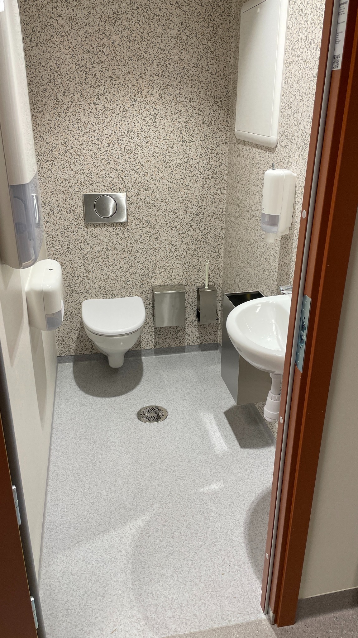 Jasna, szara posadzka w toalecie publicznej, z odpływem podłogowym. Ściany wykończone w drobną mozaikę. Widoczna umywalka, toaleta i dozowniki.