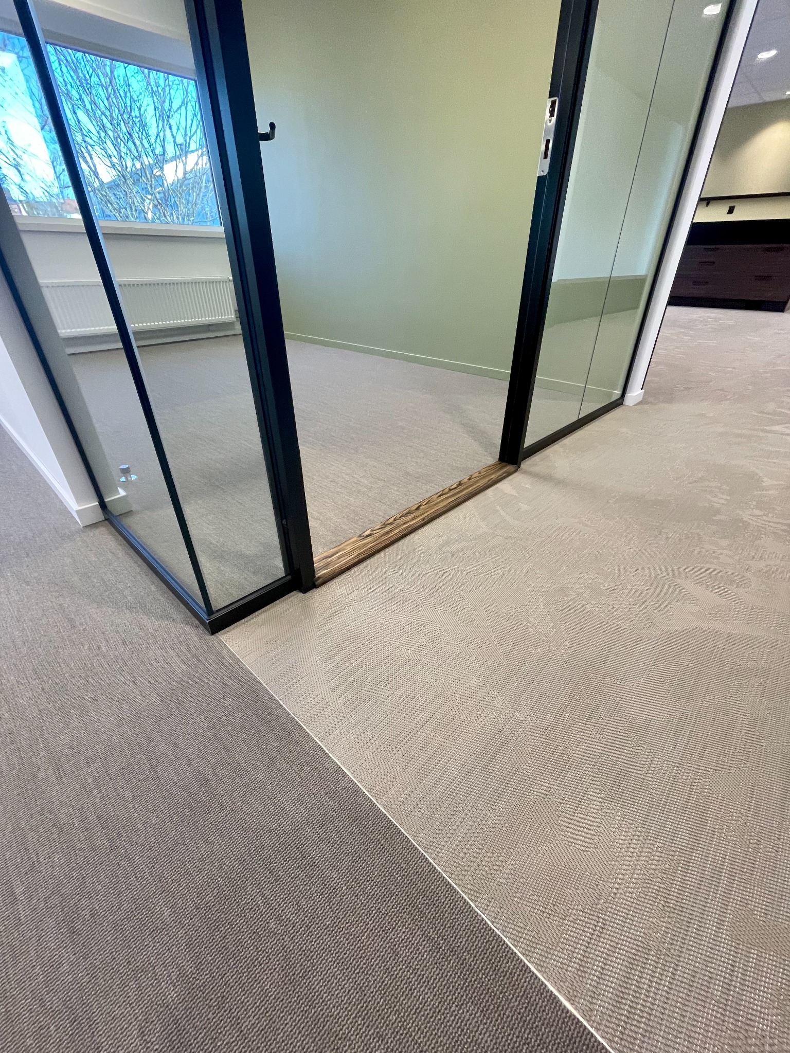 Dwie strefy biurowe z wykładziną dywanową w odcieniach szarości i beżu, oddzielone szklaną ścianą z czarną ramą i drewnianym progiem. Widok na okno i grzejnik.