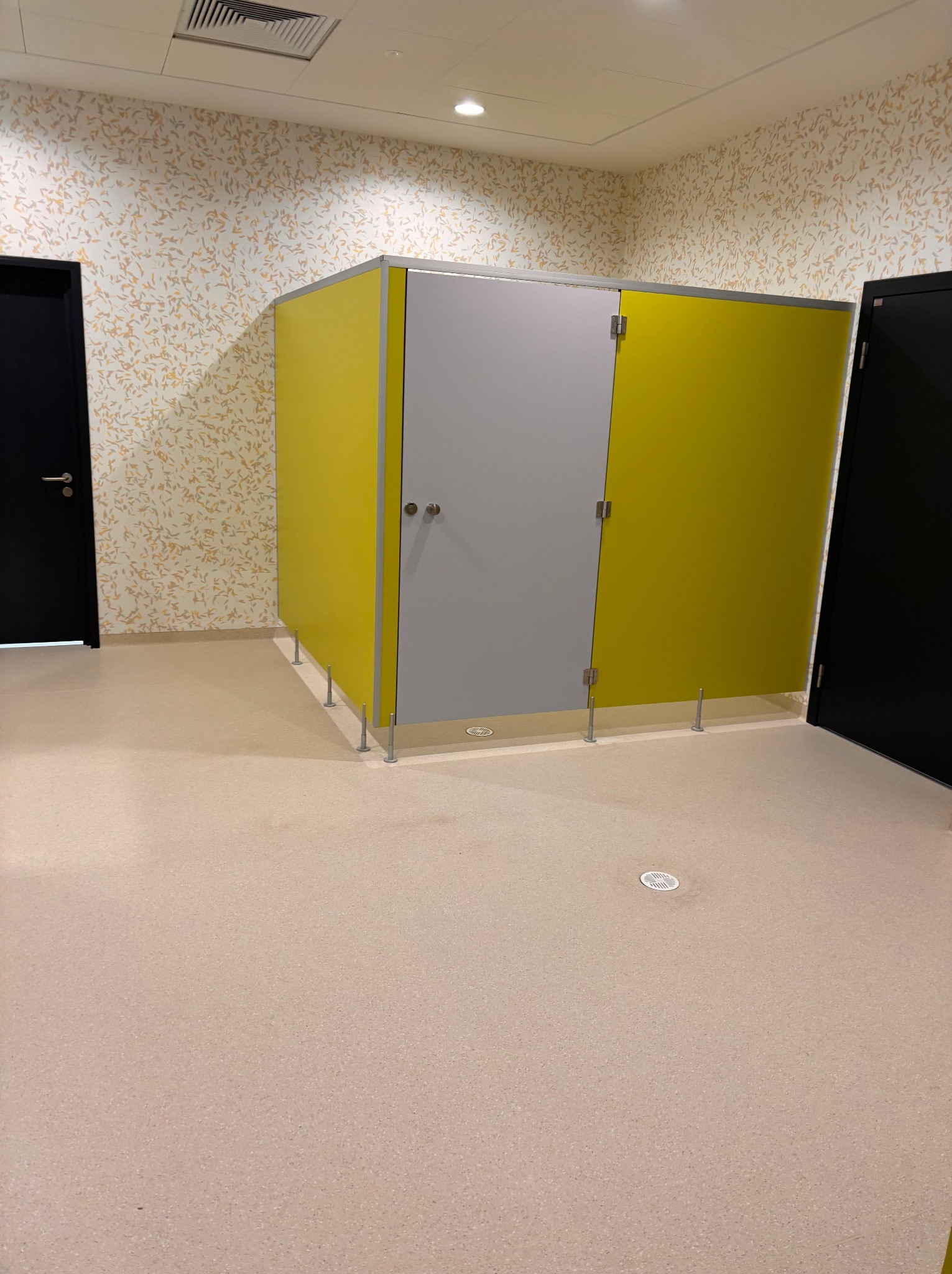 Jasna posadzka w publicznej toalecie z żółto-szarymi kabinami, widoczne detale wykończenia i wzorzysta tapeta w tle. Minimalistyczny design.