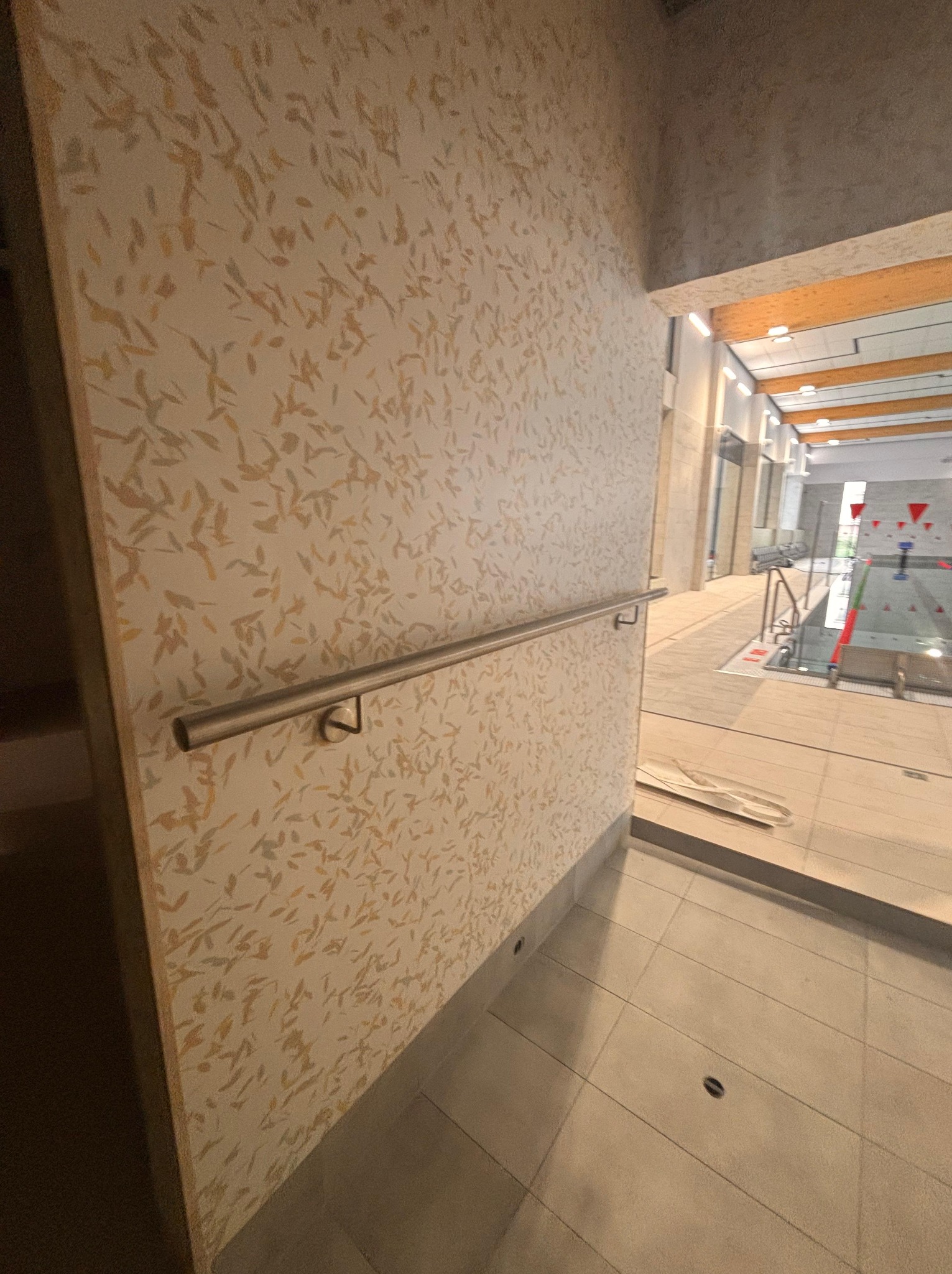 Ściana z dekoracyjną fakturą liści i metalową poręczą w hali basenowej, widok na basen z czerwonymi bojami. Jasne płytki podłogowe z punktowym oświetleniem.