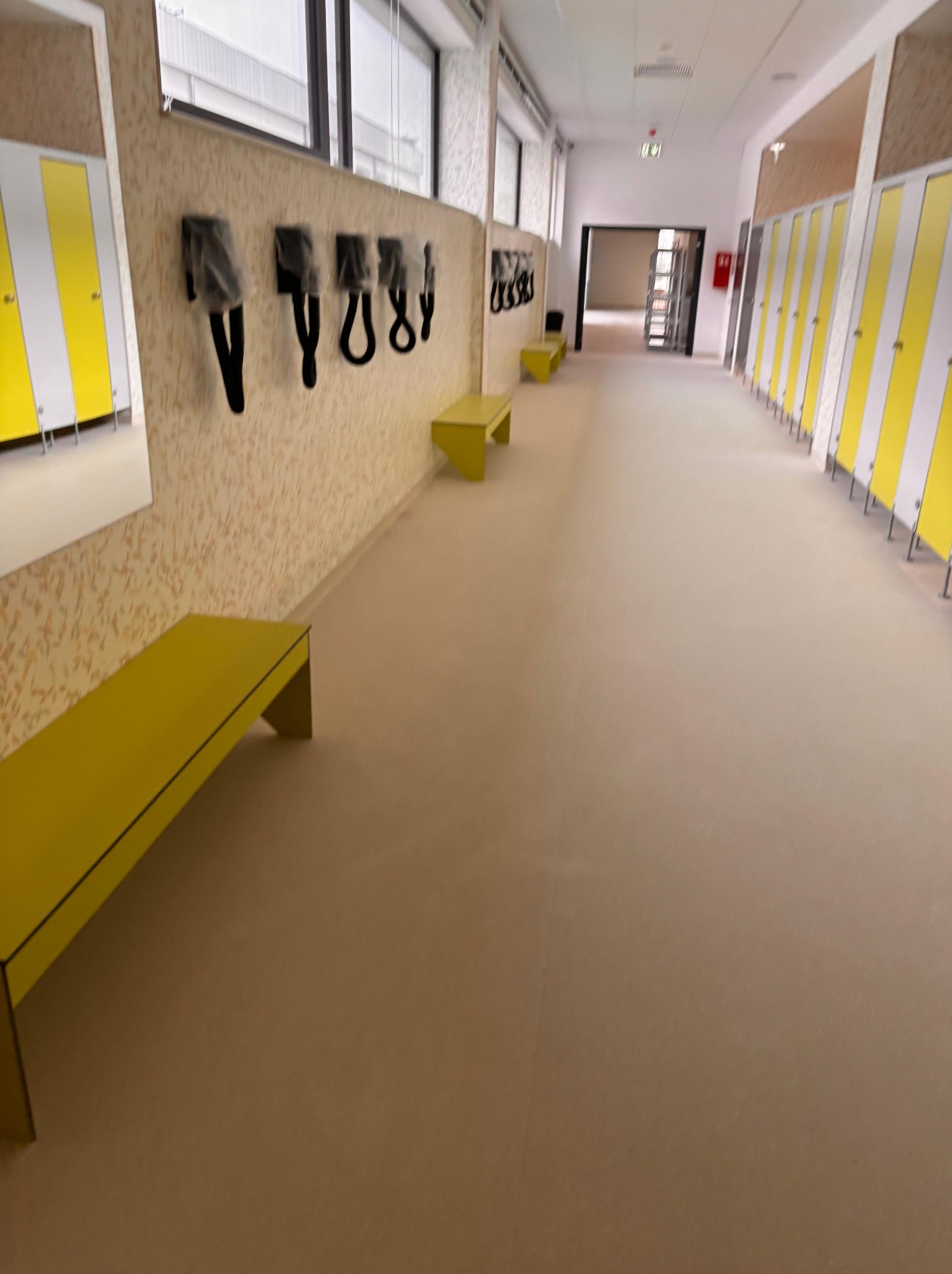 Beżowa posadzka w korytarzu szatni z żółtymi ławkami i szafkami. Na ścianie wieszaki na ubrania w folii. Widok w perspektywie zbieżnej.
