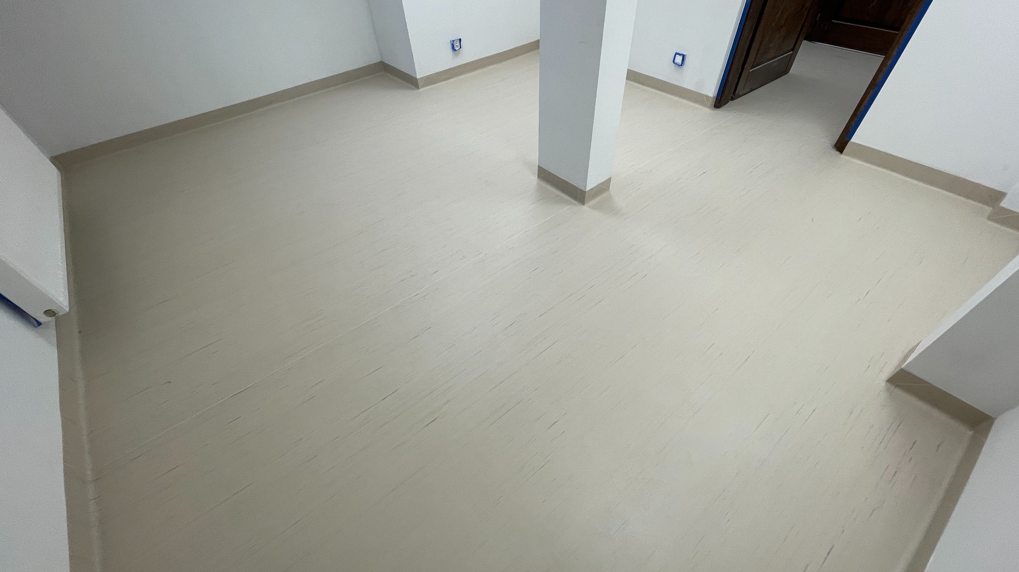 Jasna, świeżo położona wykładzina podłogowa z delikatnym wzorem, widok z góry, z kolumną w centrum pomieszczenia i otwartymi drzwiami w tle.