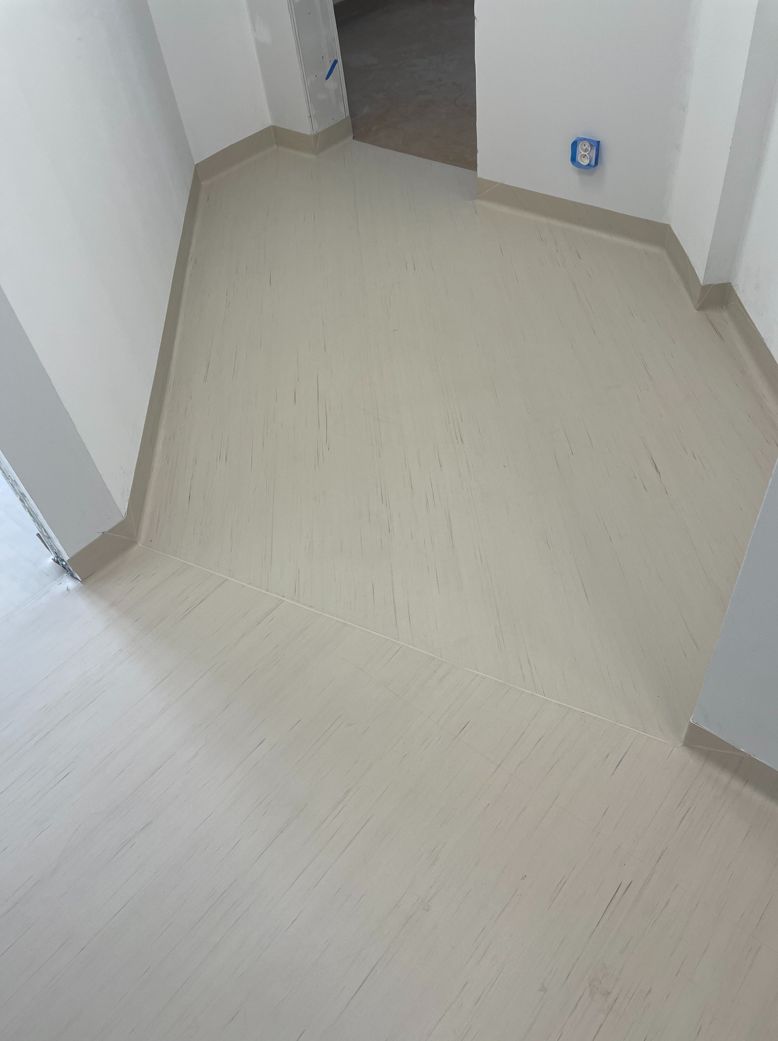 Jasna, wykończona podłoga w korytarzu z listwami przypodłogowymi. Widoczny fragment gniazdka elektrycznego na ścianie i wejście do innego pomieszczenia.