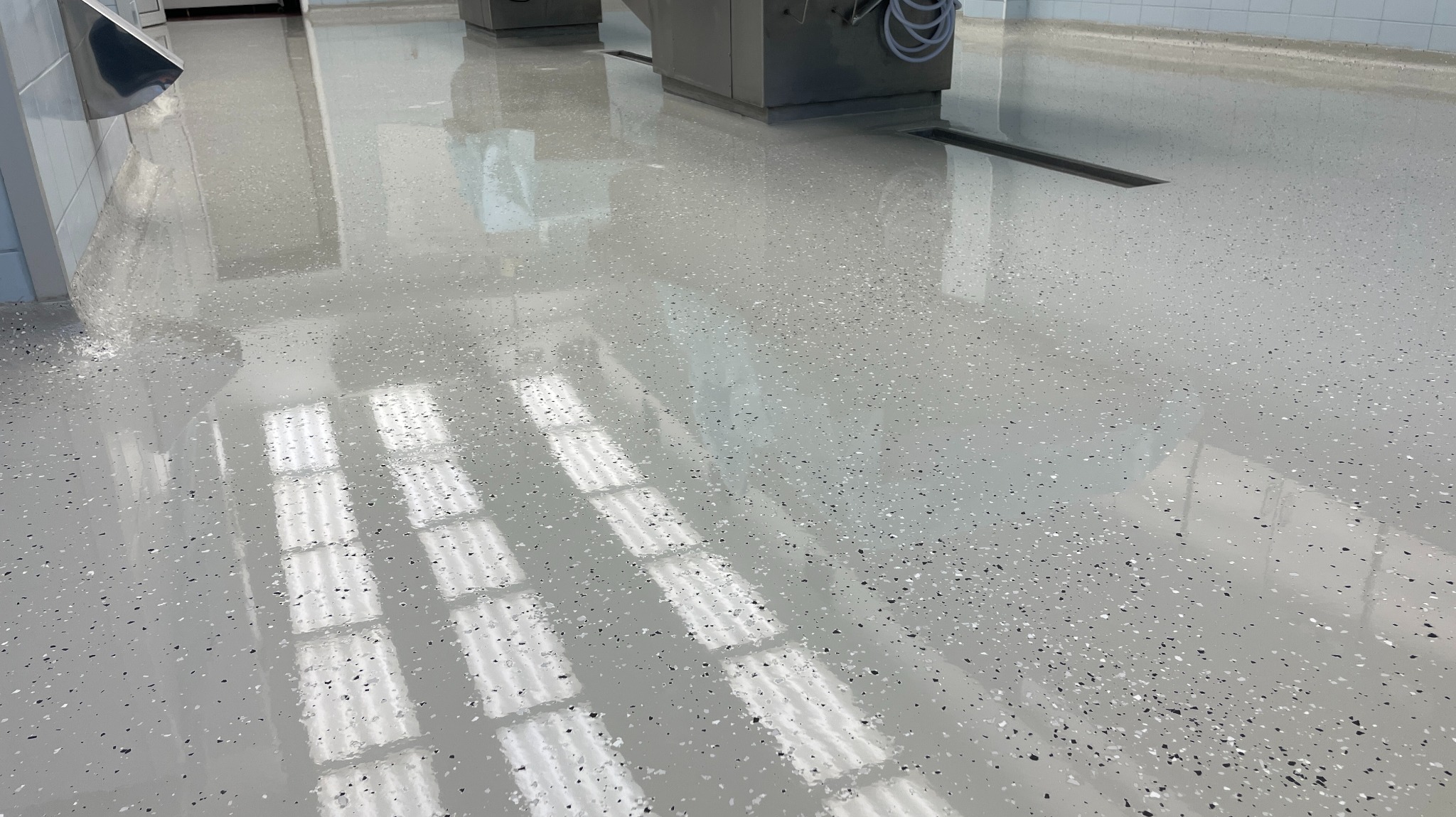 Błyszcząca, jasnoszara posadzka żywiczna z czarnymi i białymi drobinkami, odbijająca światło okien. Widoczny odpływ liniowy i fragmenty urządzeń.
