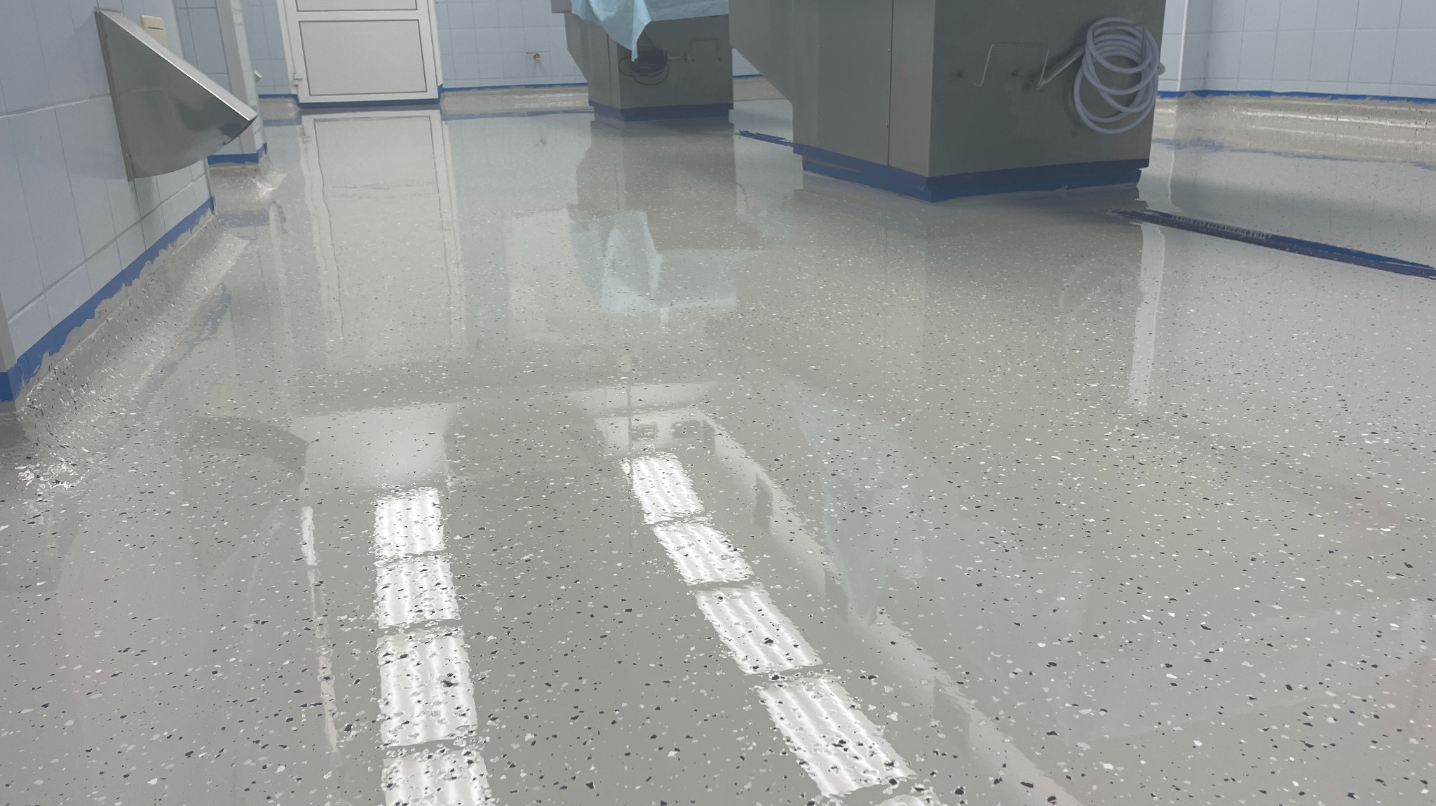 Błyszcząca, jasnoszara posadzka żywiczna z ciemnymi drobinkami w pomieszczeniu o białych kafelkach i sprzęcie medycznym. Widoczne odbicie światła w posadzce.