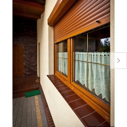 Pro Window System - Okna z drewnianą ramą i opuszczoną roletą zewnętrzną w kolorze drewna. Widoczna elewacja budynku i wejście z brązową cegłą. W oknach firanki.