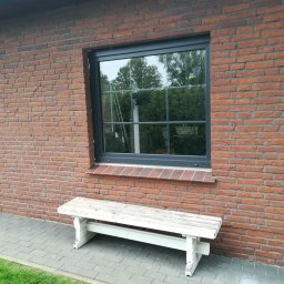 Pro Window System - Szare okno PCV z szybami dzielonymi na kwadraty, wmurowane w ścianę z czerwonej cegły. Pod oknem stoi biała, drewniana ławka. W oknie odbija się zieleń drzew.