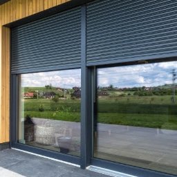 Pro Window System - Grafitowe rolety zewnętrzne, częściowo opuszczone, na oknie tarasowym z widokiem na zielony krajobraz. Nowoczesne wykończenie elewacji drewnem.