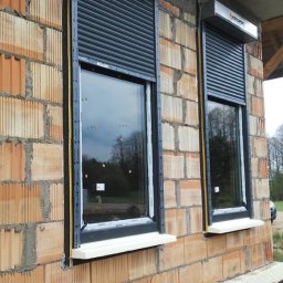 Pro Window System - Dwa okna PCV z szarymi roletami zewnętrznymi Krispol na surowej, ceglanej ścianie budynku w trakcie budowy. Widoczne ocieplenie i folia fundamentowa.