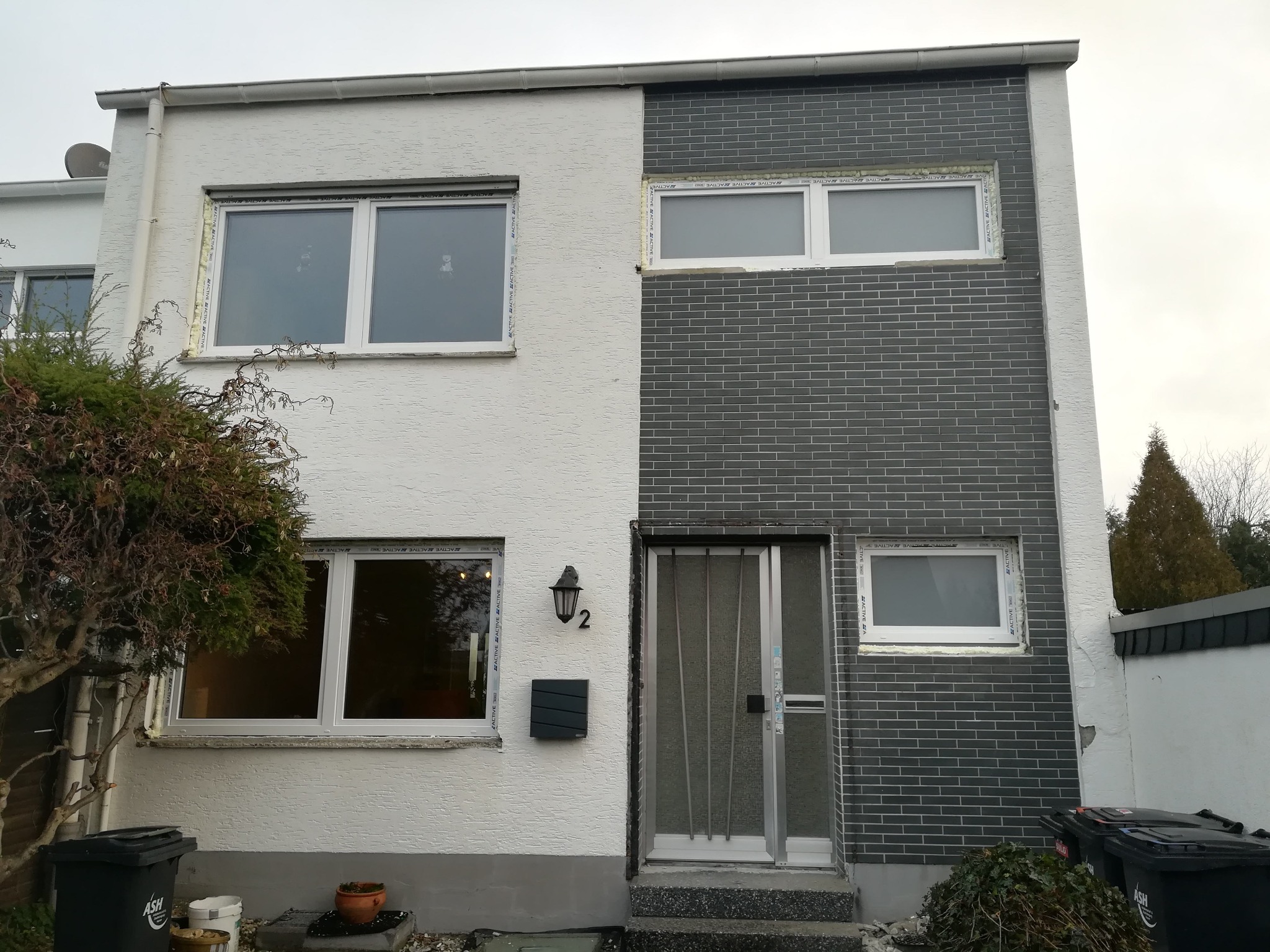 Dwukondygnacyjny dom z białymi oknami PCV i elewacją w dwóch odcieniach: białym tynku i szarej cegły. Widoczne detale wykończenia okien. Pochmurny dzień.