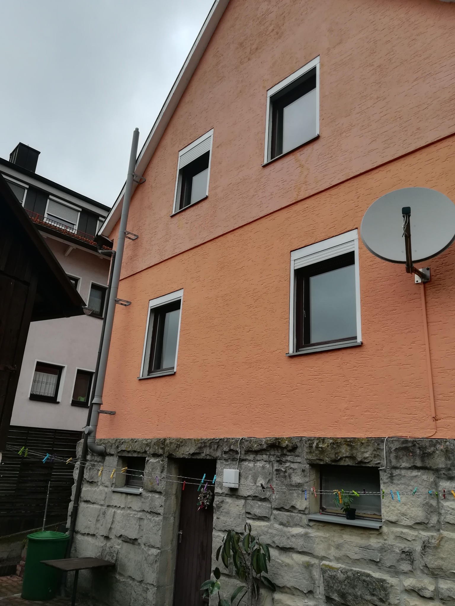 Dwukondygnacyjny dom z oknami PCV z roletami zewnętrznymi, anteną satelitarną i rynną. Dolna część budynku wykończona kamieniem.