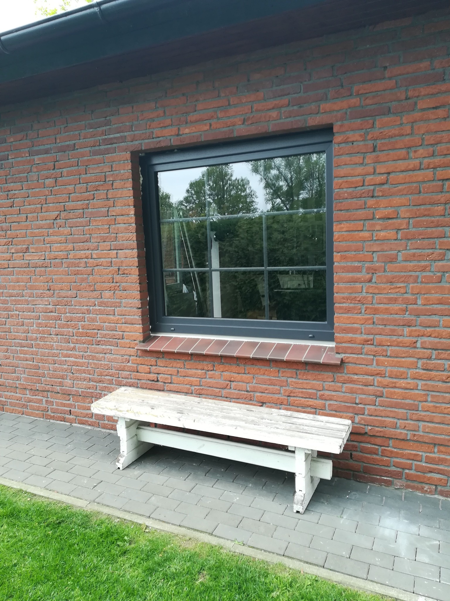 Szare okno PCV z szybami dzielonymi na kwadraty, wmurowane w ścianę z czerwonej cegły. Pod oknem stoi biała, drewniana ławka. W oknie odbija się zieleń drzew.