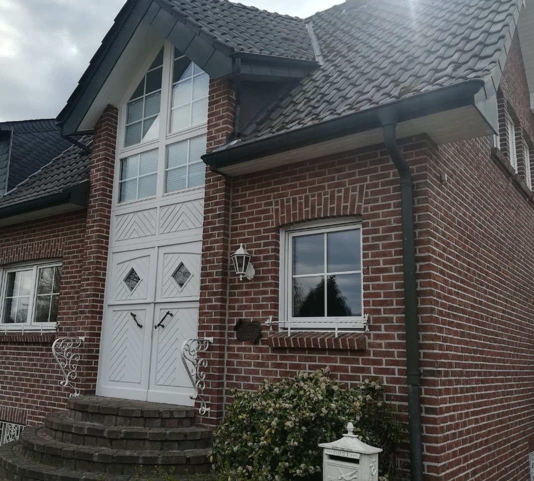 Dwuskrzydłowe, białe drzwi zewnętrzne z romboidalnymi przeszkleniami i oknem nadświetlnym w domu z czerwonej cegły. Dekoracyjne, białe balustrady.