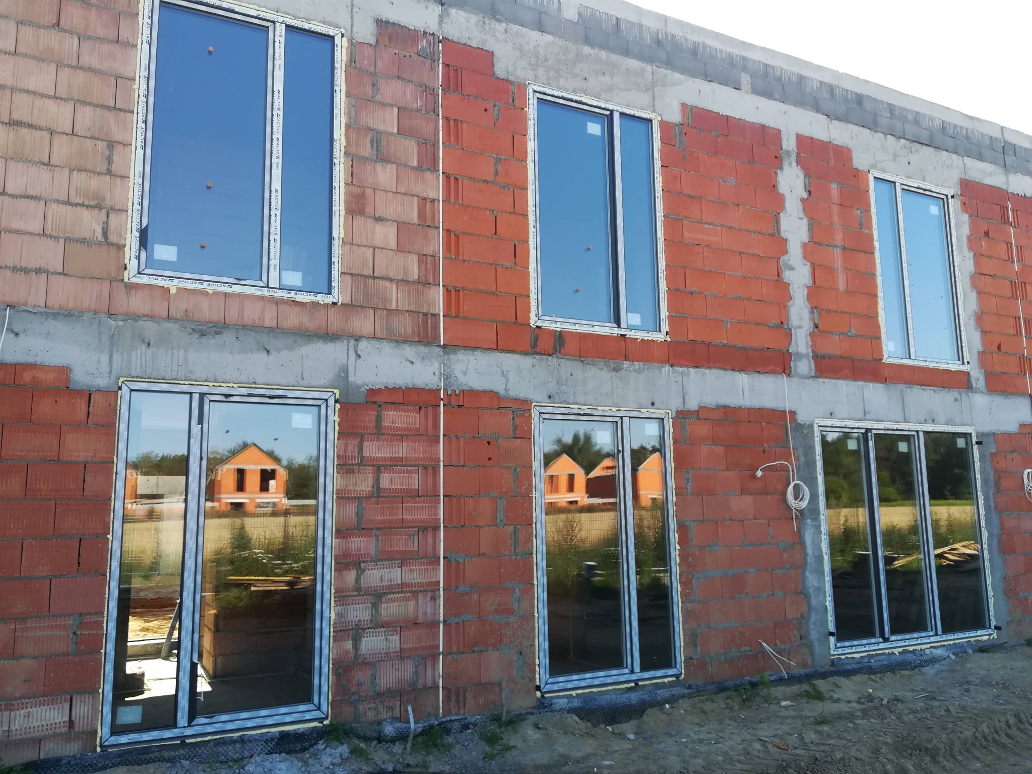 Surowy budynek z czerwonej cegły z zamontowanymi oknami PCV, w których odbija się sąsiedni dom. Widoczne szpary między oknami a murem.
