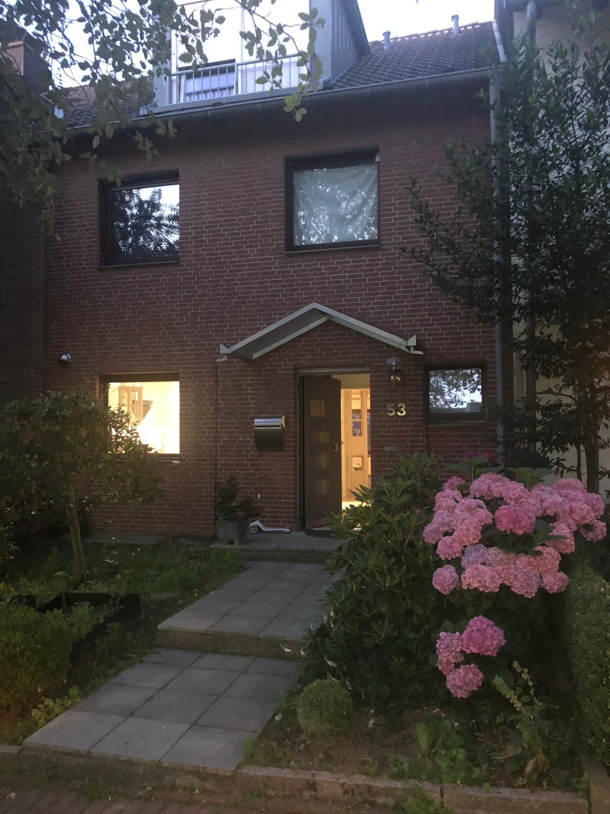 Ceglany dom z otwartymi drzwiami wejściowymi o numerze 53, widoczne okna z odbiciami. Na pierwszym planie kwitnące różowe hortensje i ścieżka z płyt.