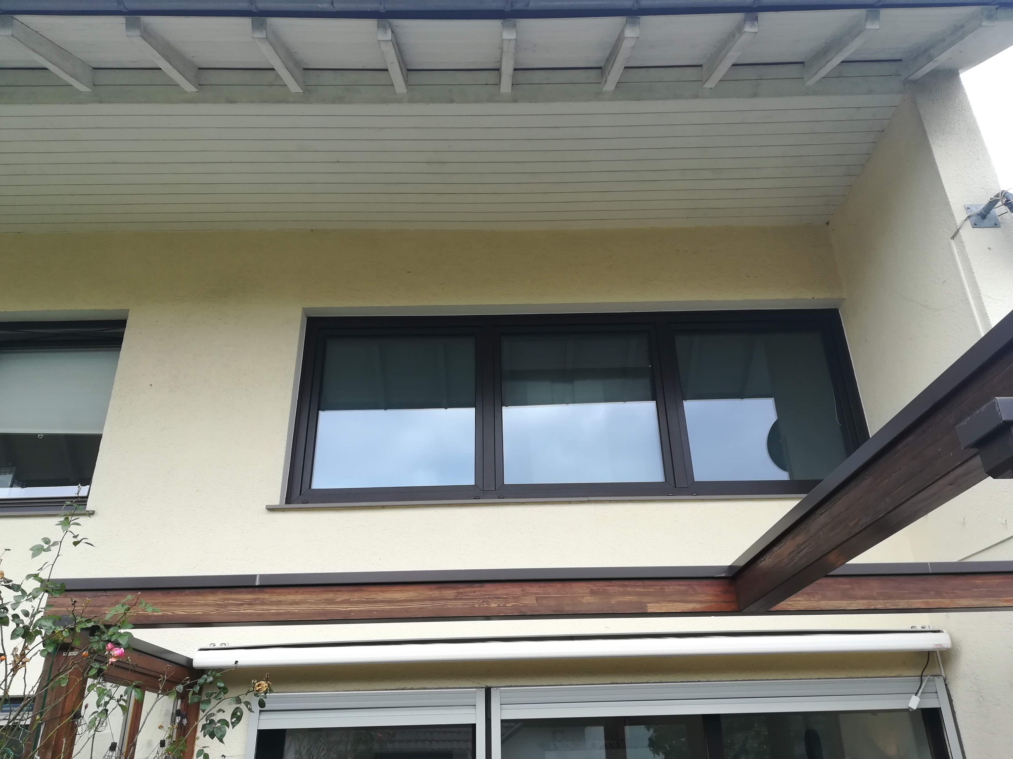 Elewacja domu z oknami PCV w ciemnych ramach, z widoczną markizą zwiniętą pod drewnianą konstrukcją. Fragment kwitnącego krzewu w lewym dolnym rogu.