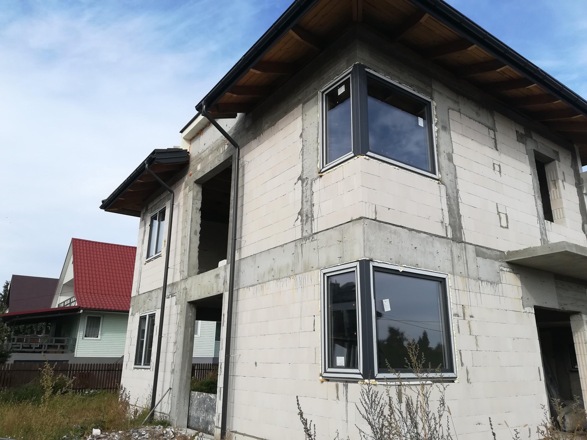 Surowy budynek w trakcie budowy z zamontowanymi oknami PCV w grafitowych ramach, widoczne bloczki i elementy konstrukcyjne, błękitne niebo w tle.