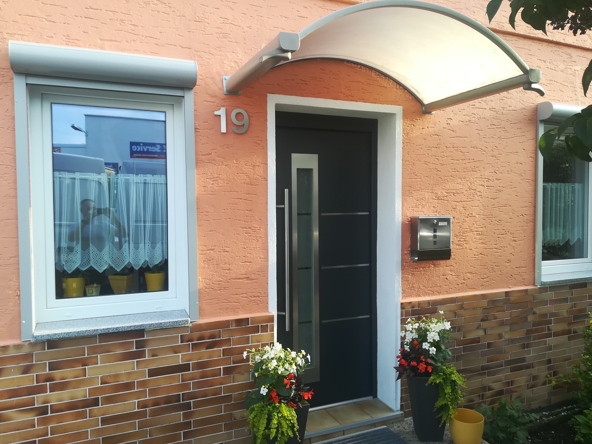Dom jednorodzinny z oknem z roletą i nowoczesnymi drzwiami wejściowymi pod zadaszeniem, z numerem 19. Dekoracyjne kwiaty w donicach po bokach wejścia.
