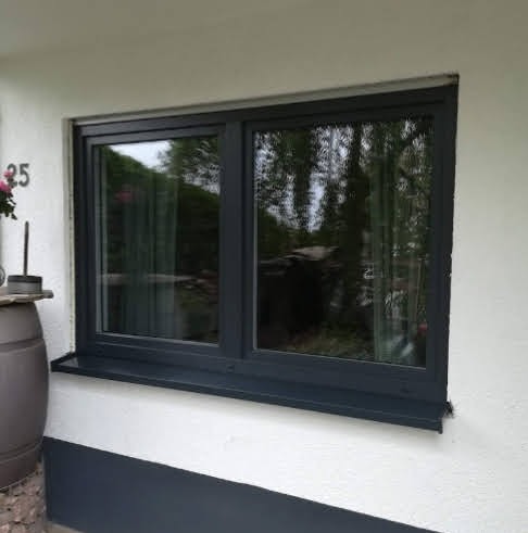 Zamontowane okno PCV w grafitowej ramie, z widocznym odbiciem zieleni ogrodu. Dolna część ściany pomalowana na szaro, numer domu 25 z lewej strony.