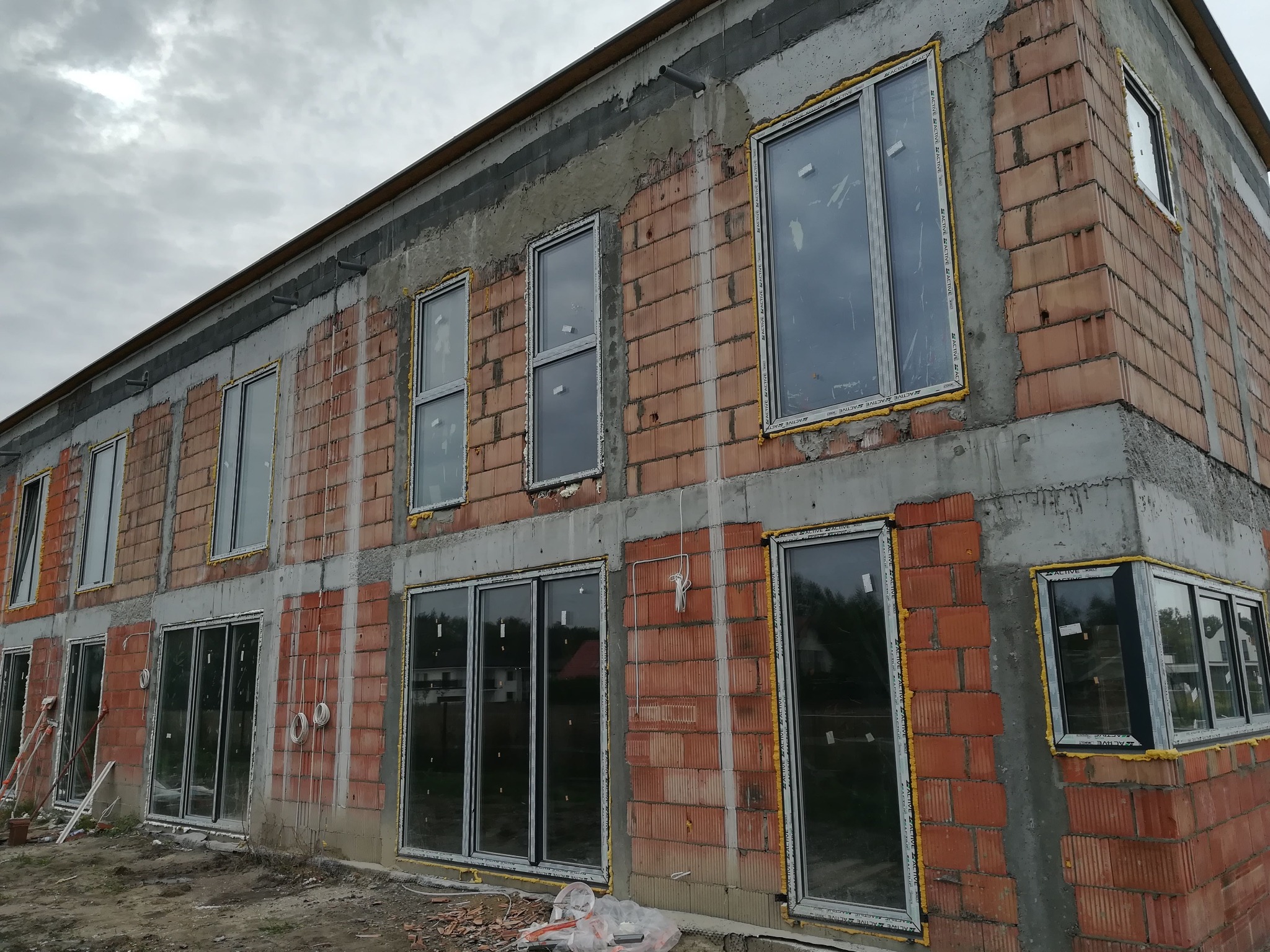 Surowy stan budynku w trakcie montażu okien PCV, widoczne okna z pianką montażową. Budynek z czerwonej cegły, częściowo otynkowany, pochmurne niebo w tle.