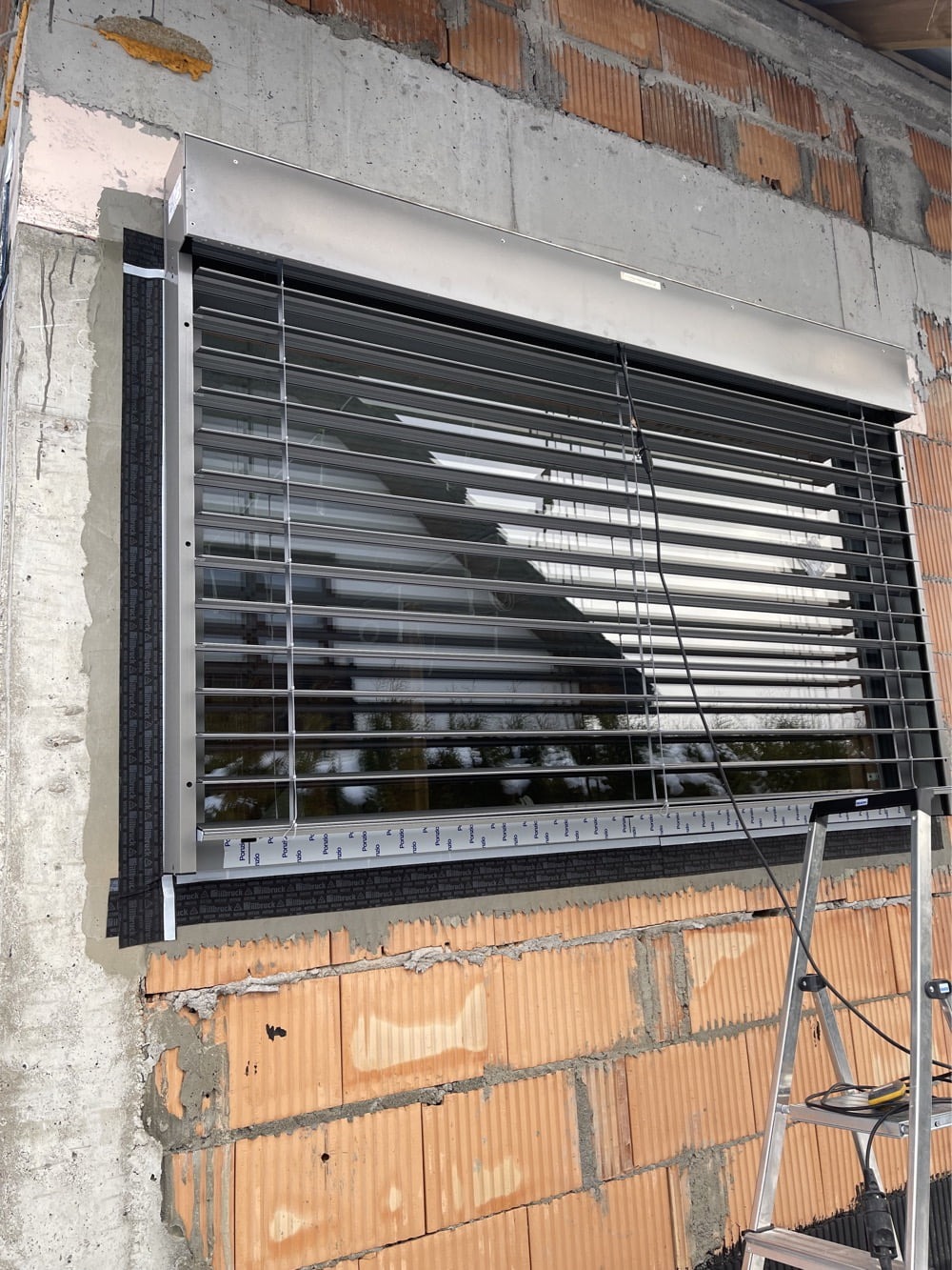 Szara roleta antywłamaniowa zamontowana na oknie budynku w trakcie budowy. Widoczna drabina i narzędzia. Surowy mur z cegły i betonu.
