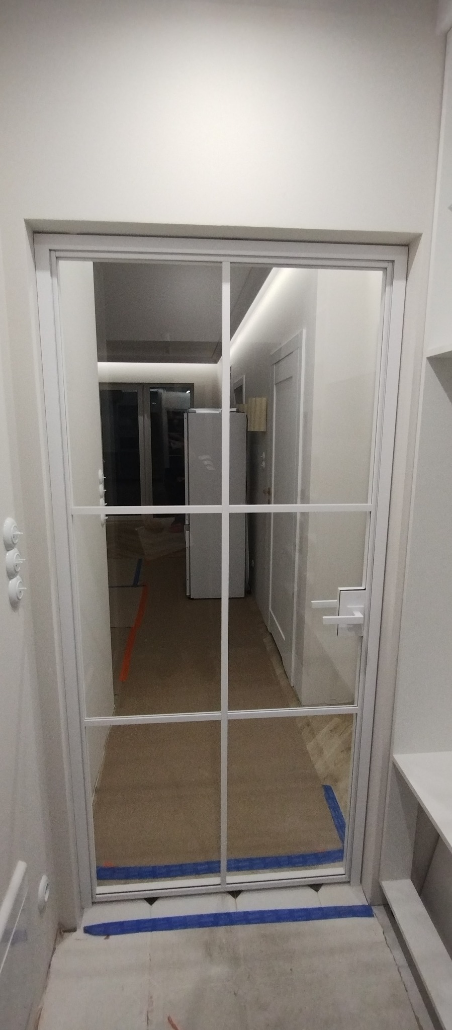 Białe, przeszklone drzwi wewnętrzne z kratownicą, widok na korytarz z białymi ścianami i oświetleniem LED. Podłoga zabezpieczona folią malarską.