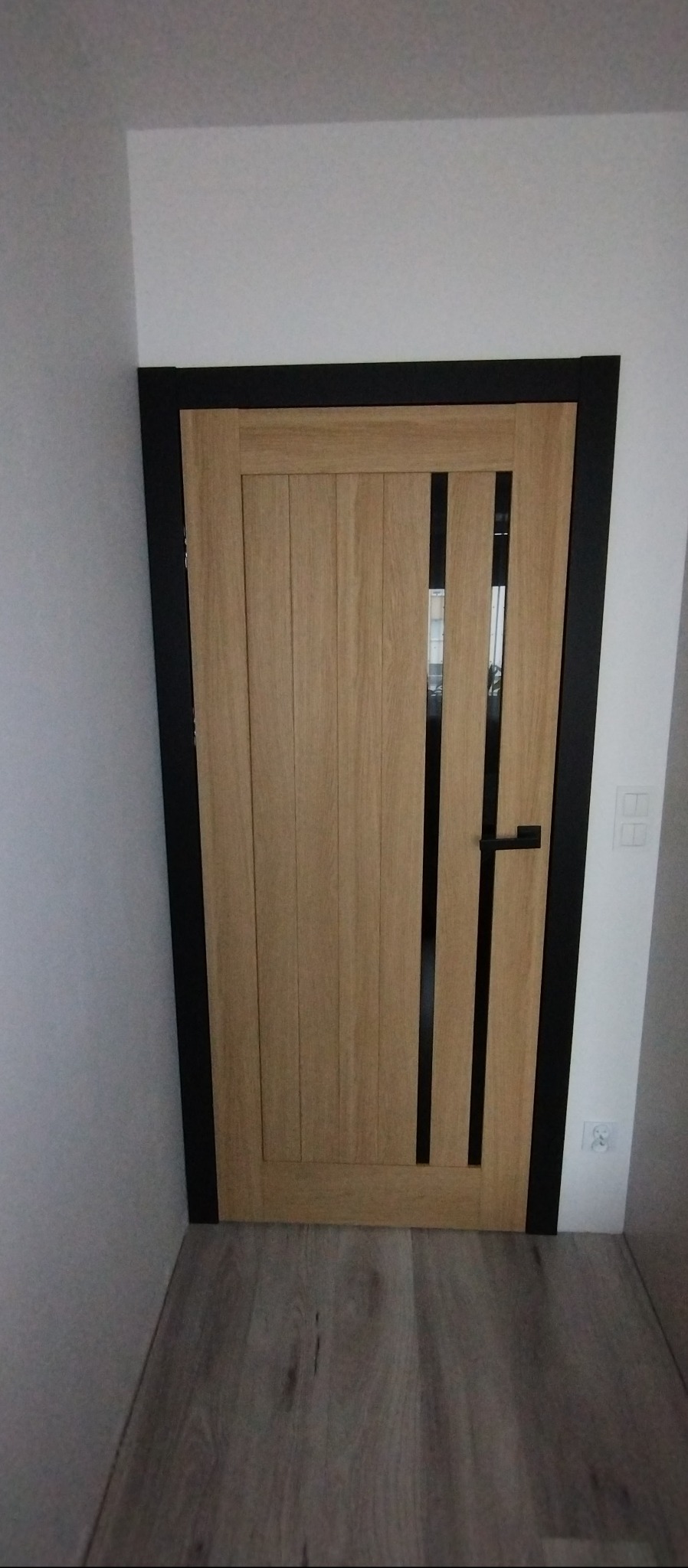 Dębowa okleina drzwi wewnętrznych z czarną ramą i trzema pionowymi intarsjami, czarna klamka, widok na fragment podłogi i ściany z włącznikiem.