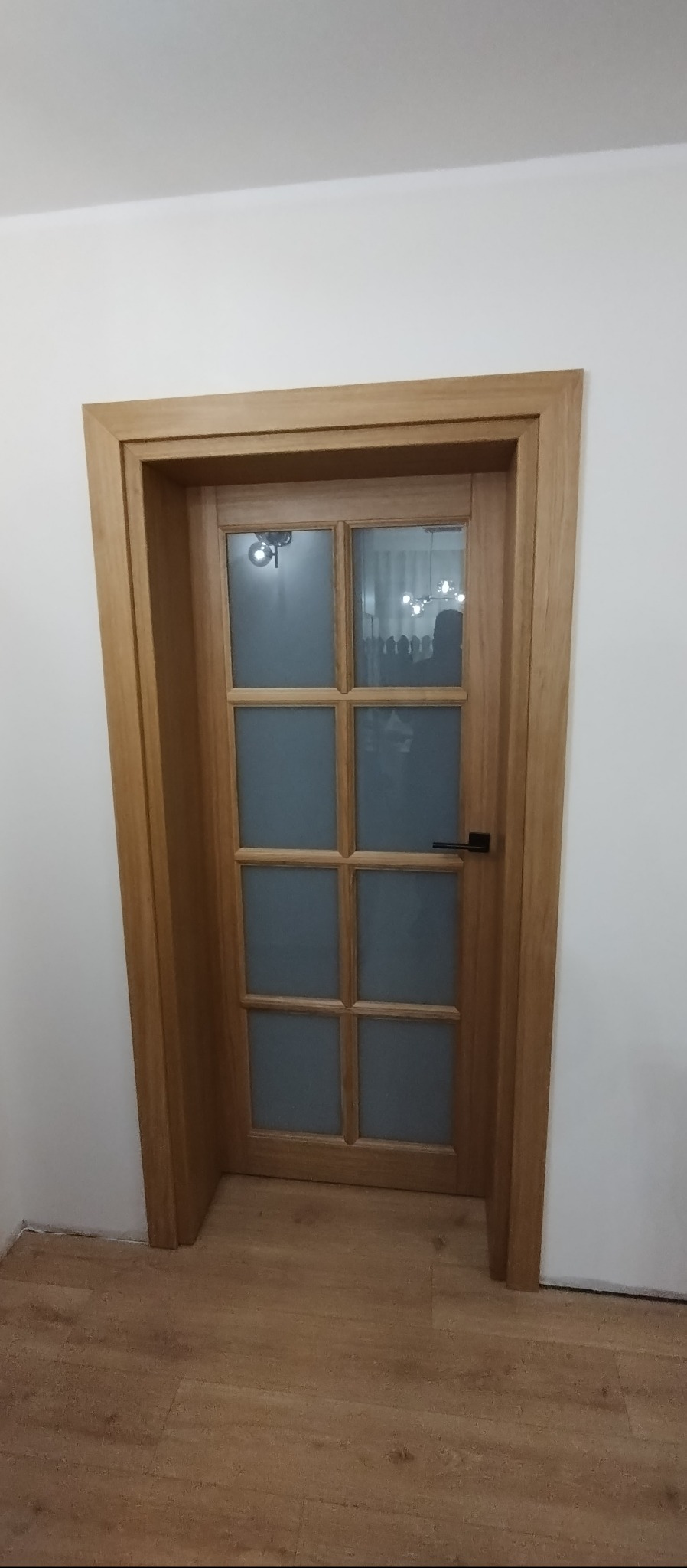 Drewniane drzwi wewnętrzne z szybami w ramie, czarna klamka, widok na wnętrze z żyrandolem. Naturalne oświetlenie, parkiet na podłodze, białe ściany.