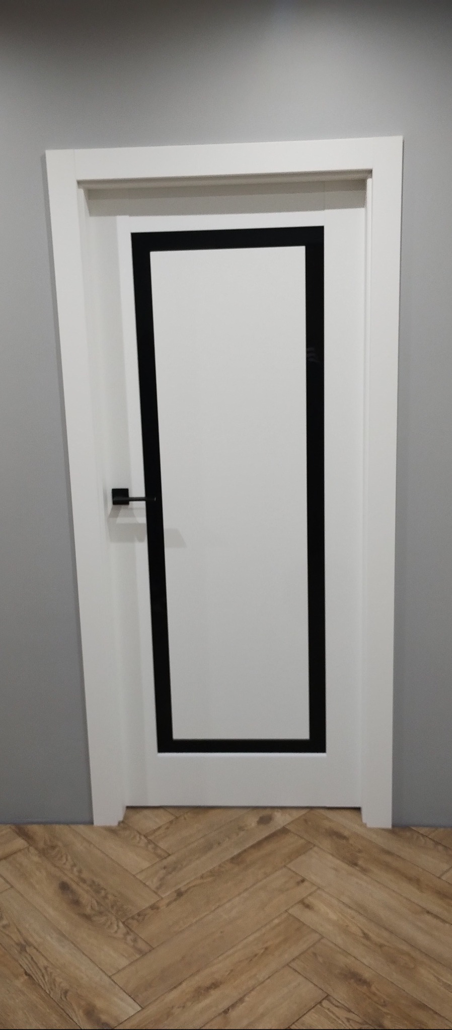 Białe drzwi wewnętrzne z czarną, prostokątną ramką na tle szarej ściany i podłogi z paneli ułożonych w jodełkę. Minimalistyczny design z czarną klamką.