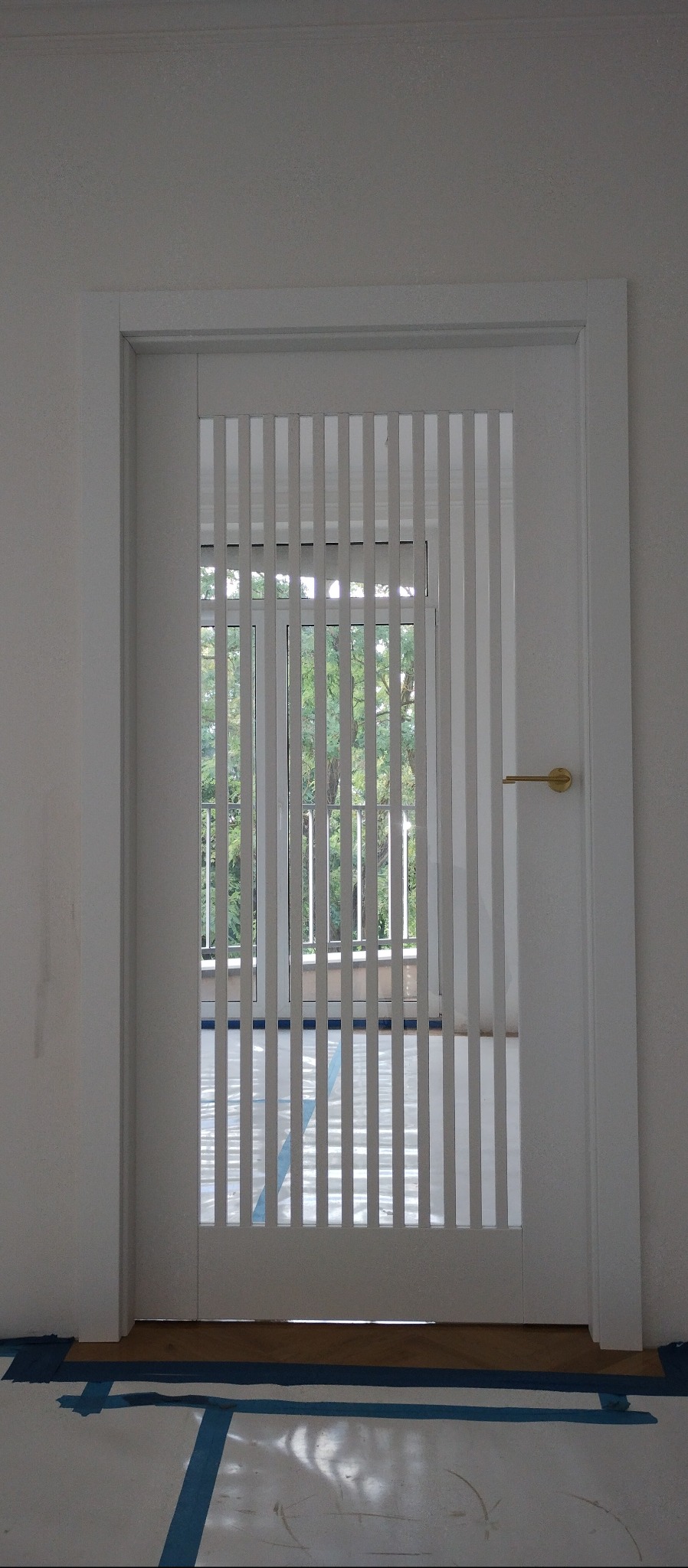 Białe drzwi wewnętrzne z pionowymi listwami w Białystoku, przez które widać okno i zieleń. Złota klamka po prawej stronie. Podłoga zabezpieczona taśmą.