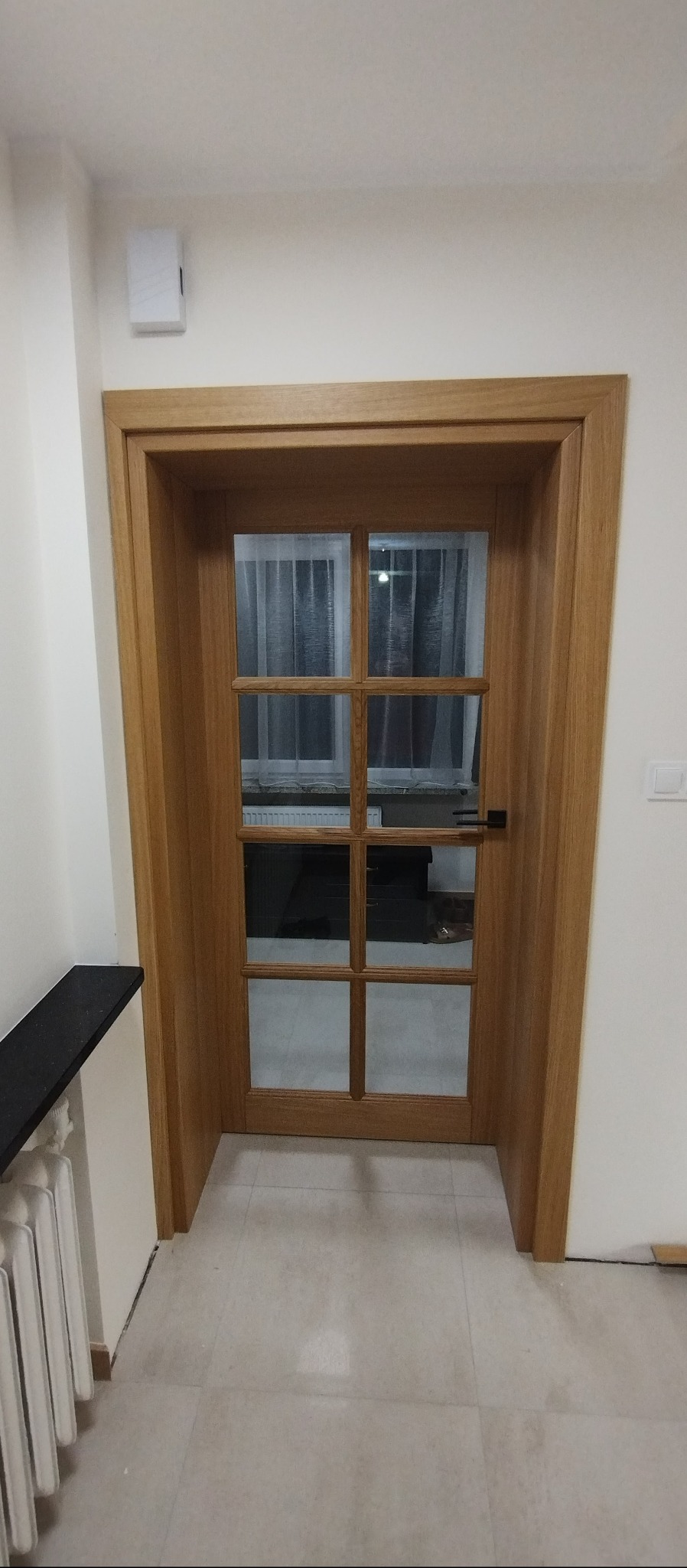 Drewniane drzwi wewnętrzne z szybami w szprosach, czarna klamka, jasna podłoga. Widok na wnętrze przez szyby. Neutralne oświetlenie.