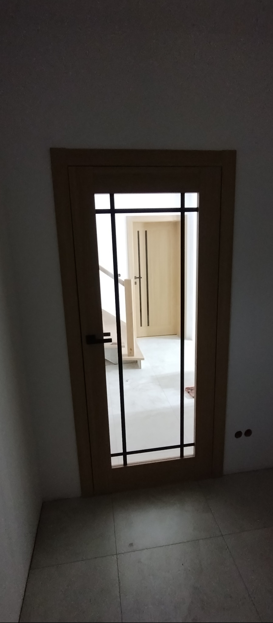 Beżowe drzwi wewnętrzne z czarnymi szprosami, widok na klatkę schodową i kolejne drzwi. Minimalistyczny design, jasne wnętrze, płytki podłogowe w odcieniach szarości.
