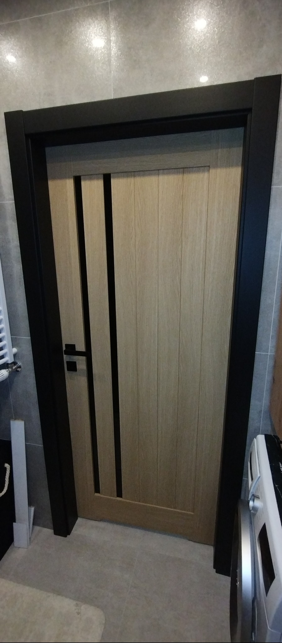 Jasne drzwi wewnętrzne z czarnymi, pionowymi wstawkami i czarną ościeżnicą w łazience z szarymi płytkami. Minimalistyczny design.