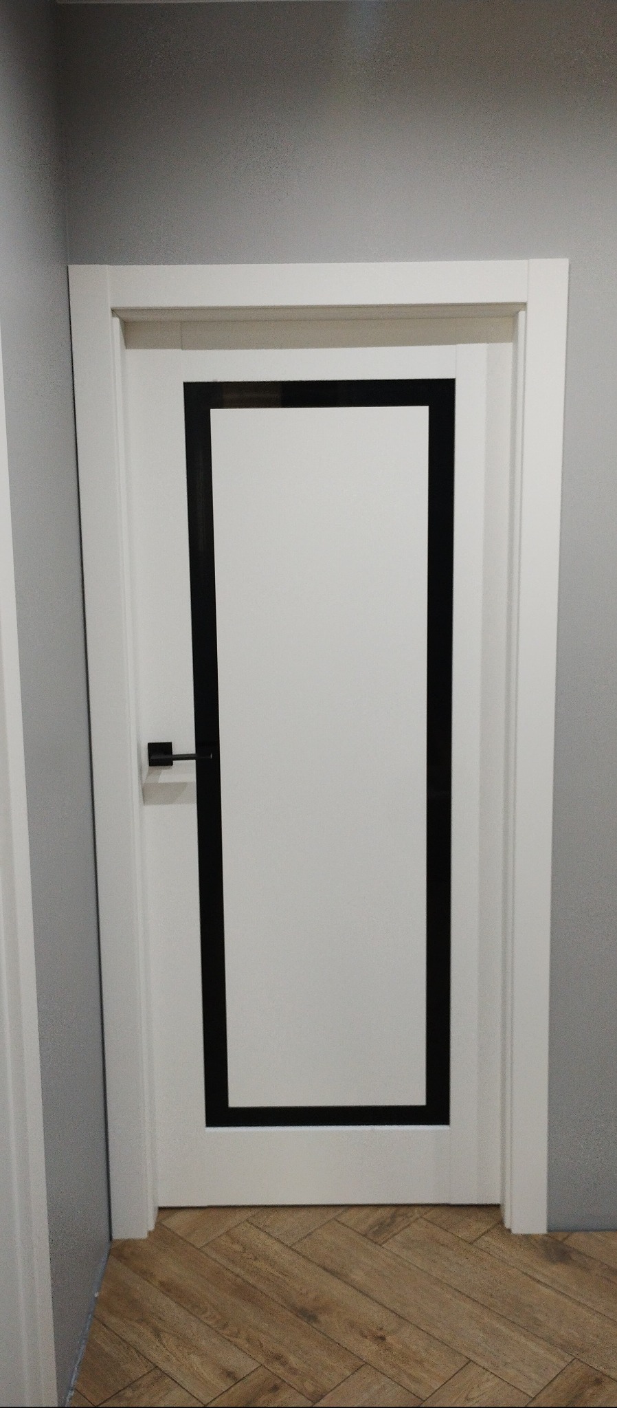 Białe drzwi wewnętrzne z czarną, prostokątną ramką, zamontowane w szarej ścianie. Widoczna klamka i podłoga z paneli ułożonych w jodełkę.