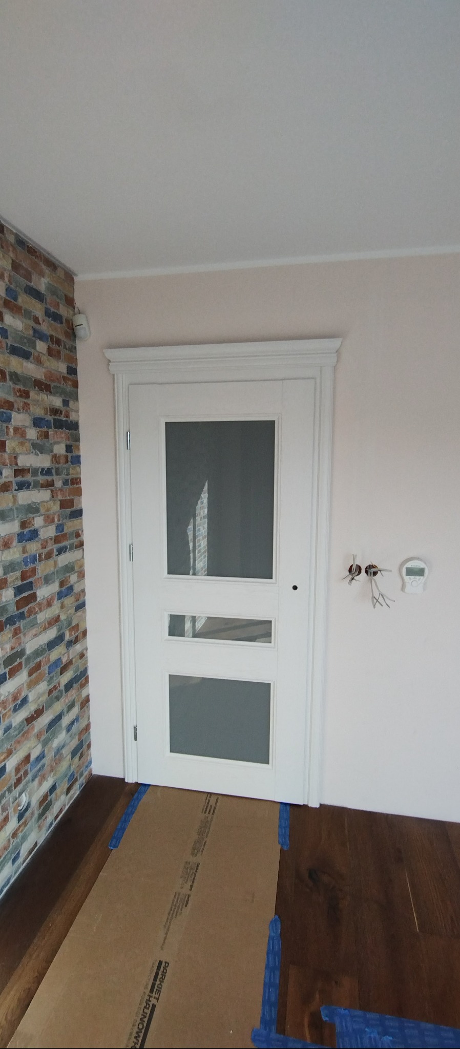 Białe drzwi wewnętrzne z trzema szybami, widoczne na tle ściany z cegły i jasnej ściany z wystającymi kablami. Podłoga zabezpieczona kartonem.