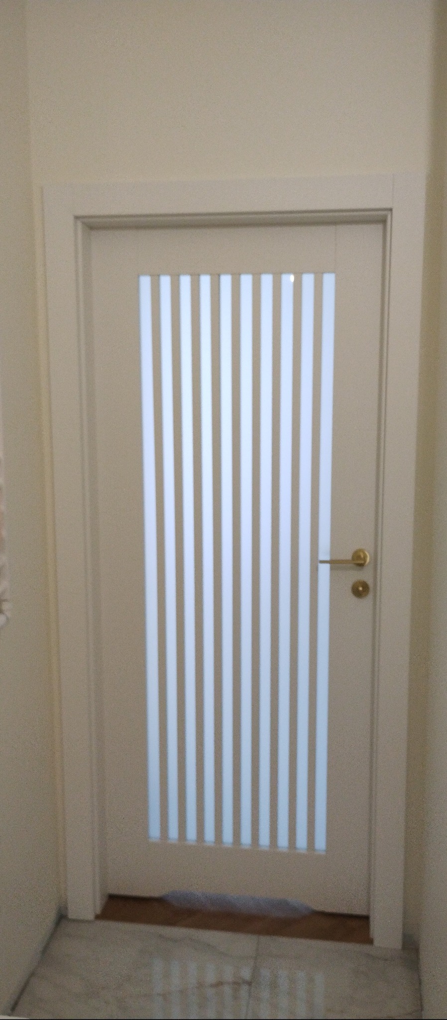 Białe drzwi wewnętrzne z szybą w pionowe paski i złotą klamką, zamontowane w jasnym pomieszczeniu. Widoczny fragment podłogi.