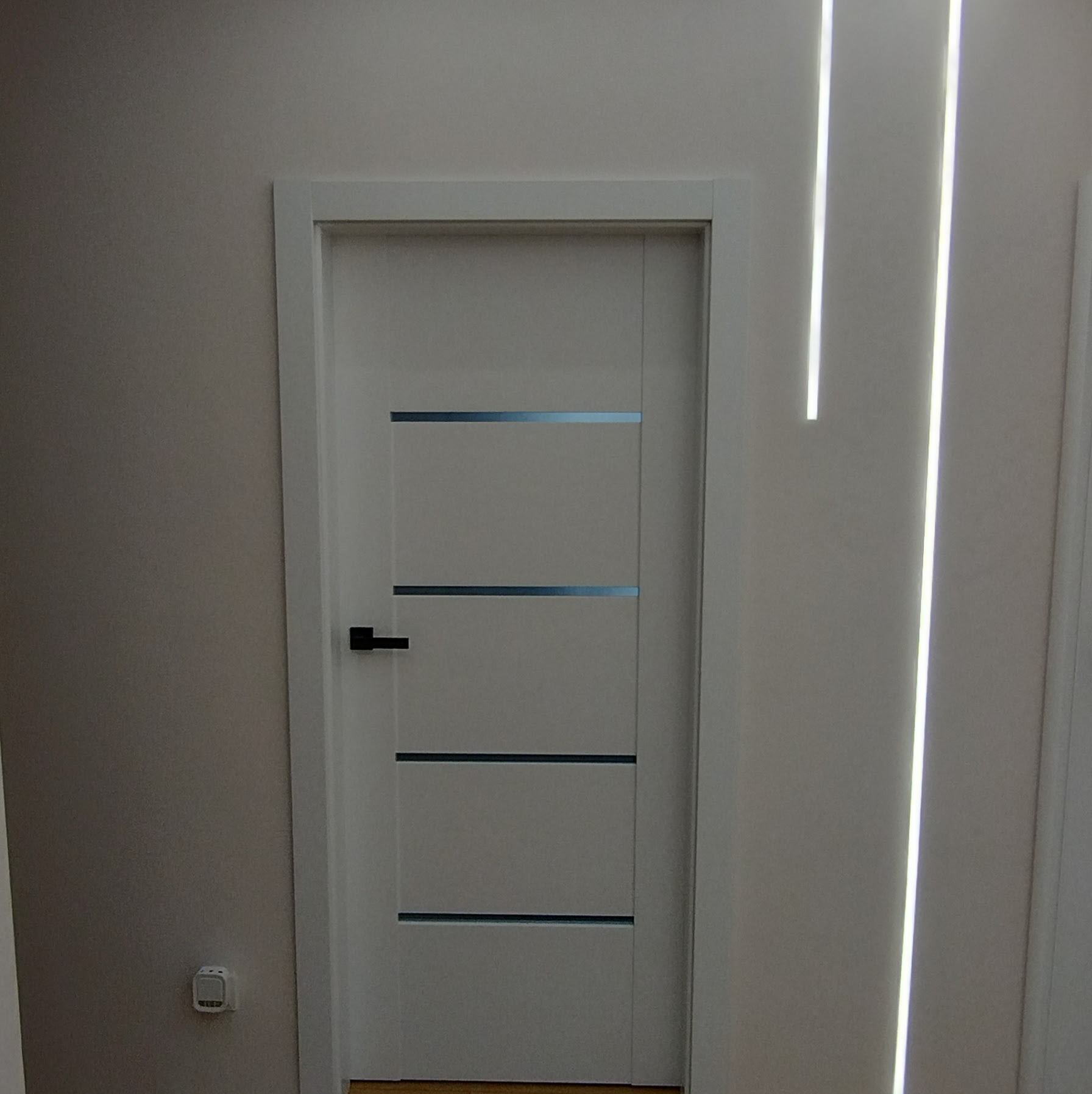Białe drzwi wewnętrzne z czarną klamką i poziomymi, szarymi wstawkami, w białej futrynie. Na ścianie pionowe, ledowe oświetlenie.