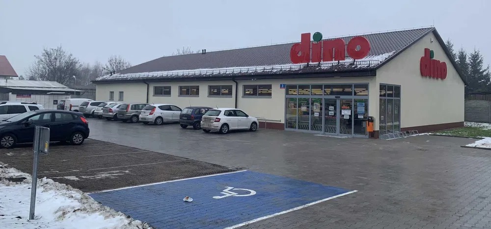 Parking przed sklepem Dino w pochmurny dzień. Widoczna kostka brukowa, zaparkowane samochody i fragmenty topniejącego śniegu.