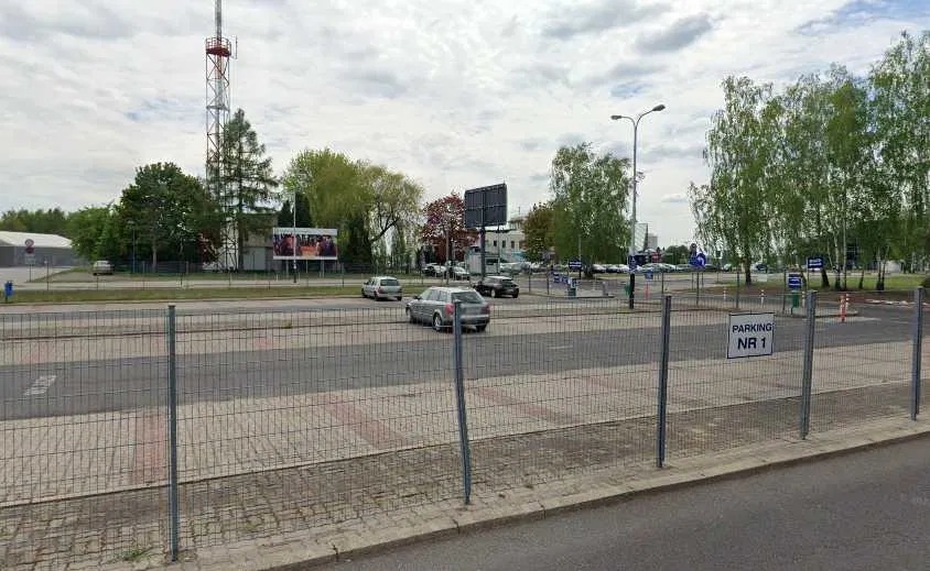 Ogrodzenie z siatki na tle parkingu z samochodami i zielenią miejską w pochmurny dzień. Widoczny znak 'Parking Nr 1'.