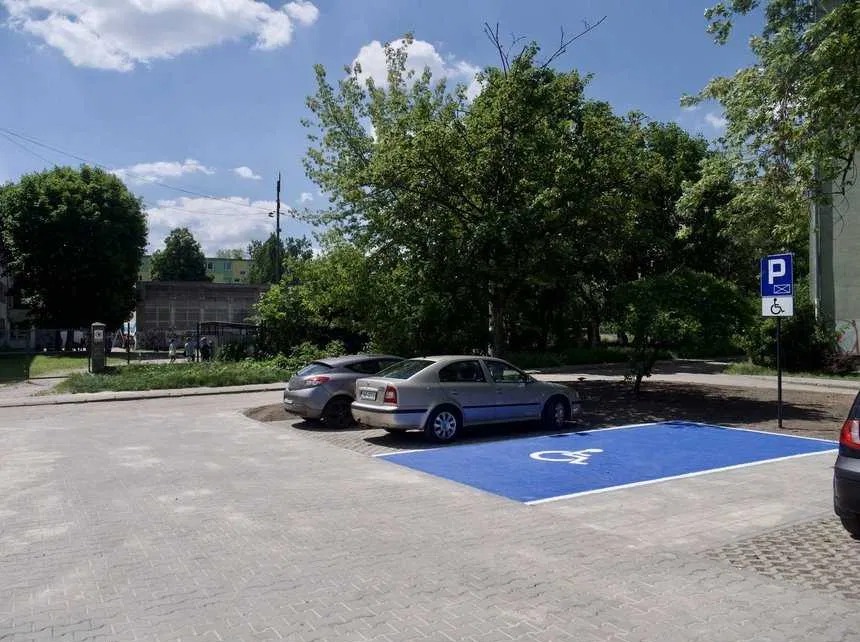 Szara kostka brukowa z niebieskim miejscem parkingowym dla niepełnosprawnych, znak pionowy, dwa zaparkowane auta i drzewa w tle, słoneczny dzień.