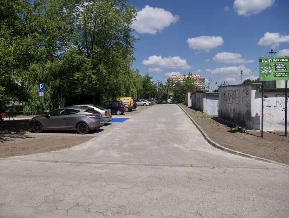 Ukończony parking z kostki brukowej, z zaparkowanymi samochodami. Widoczny znak parkingowy i budynki w tle.