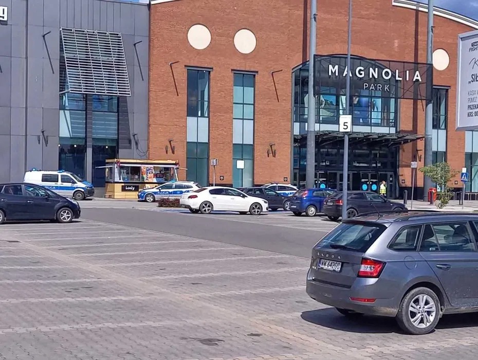Parking przed Magnolia Park we Wrocławiu, z szarą Skodą Fabią na pierwszym planie. W tle widoczne radiowozy policyjne i budka z lodami.
